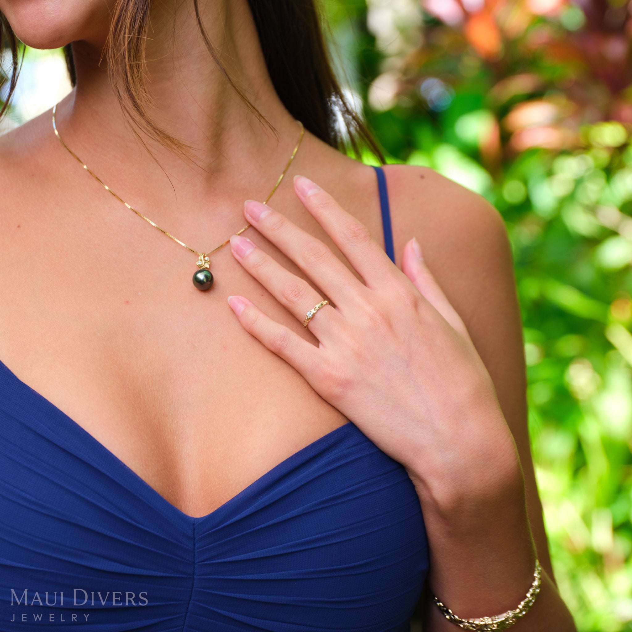 Woman in a blue top wearing a Living Heirloom Ring in 14k yellow gold with a diamond on her ring finger, with her hand placed next to a Tahitian black pearl pendant around her neck, against a blurred tropical background