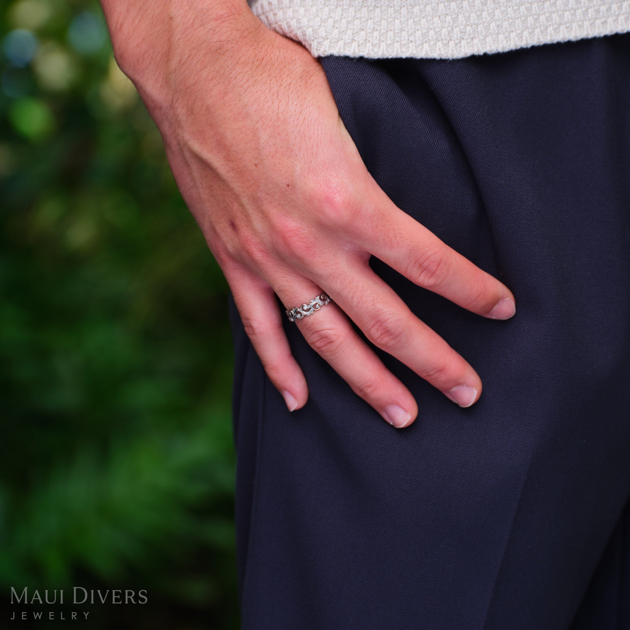 Close-up of a Living Heirloom Ring in 14k white gold with diamond accents worn on the ring finger, with a hand in the pocket of dark blue pants, against a blurred green background