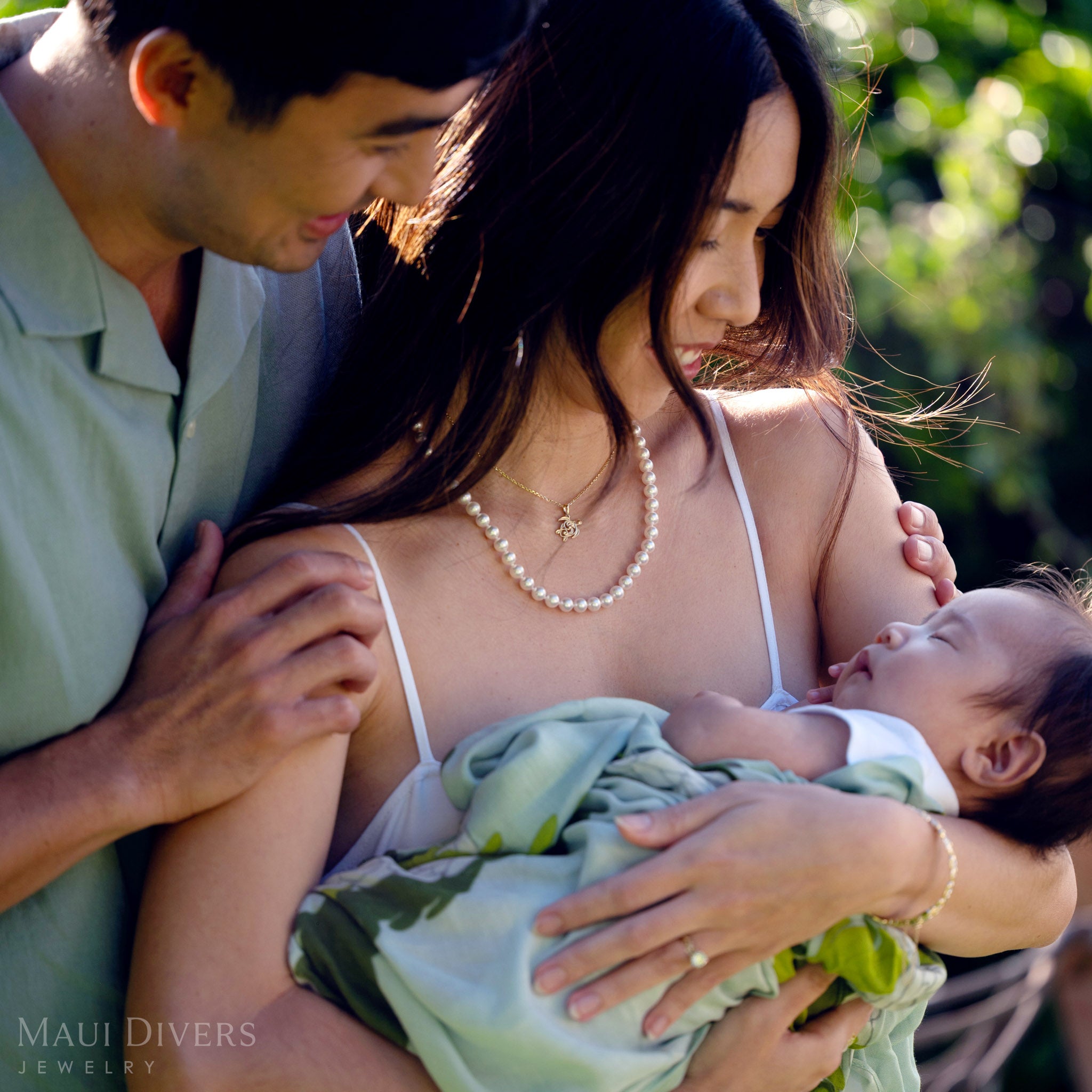 Smiling woman wearing a Living Heirloom Honu pendant in 14k yellow gold and a white pearl strand necklace, looking at a baby with a man smiling over her shoulder, against a blurred green background