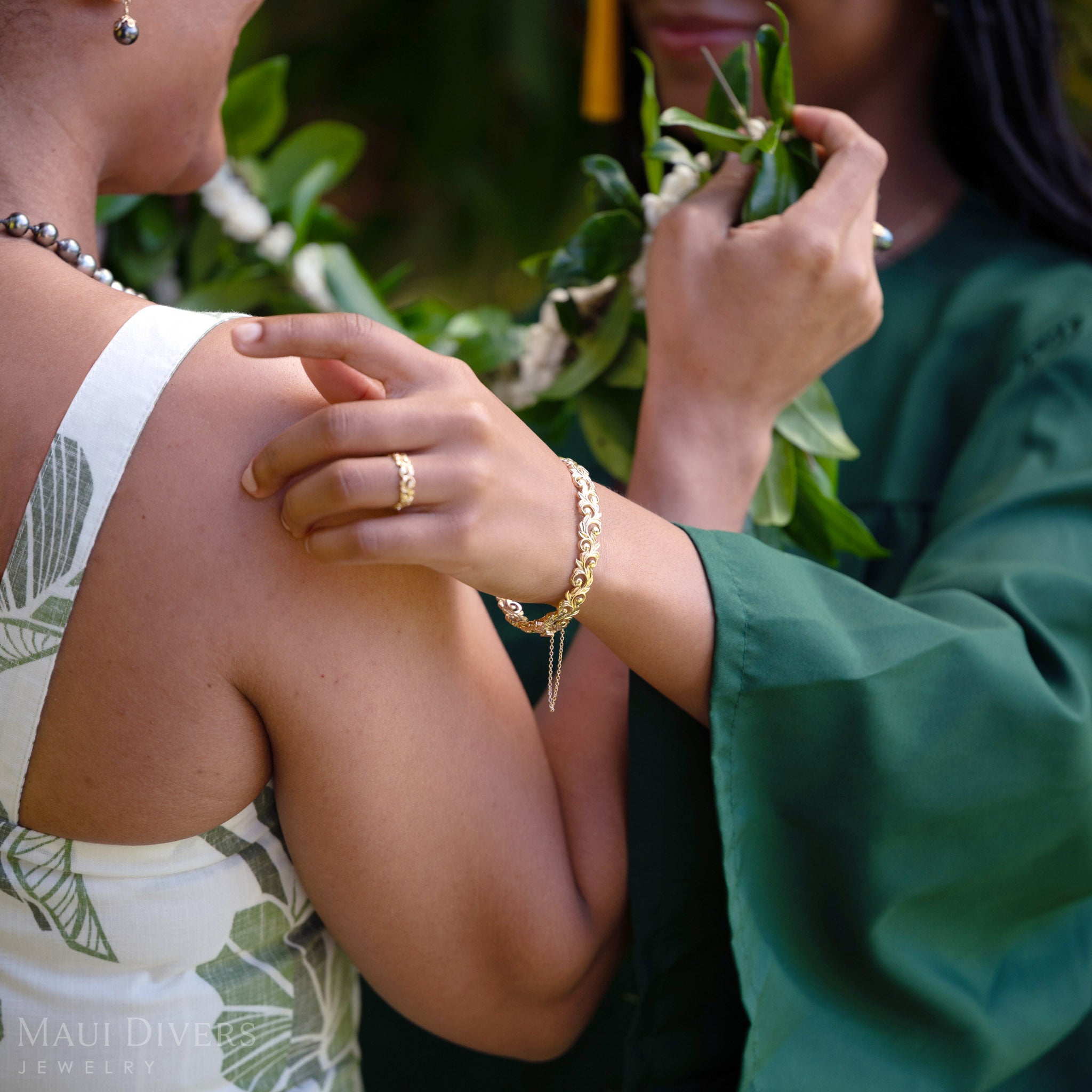 Close-up of a Living Heirloom Hinge Bracelet in 14k yellow gold worn on the left wrist, facing another woman against a blurred green background