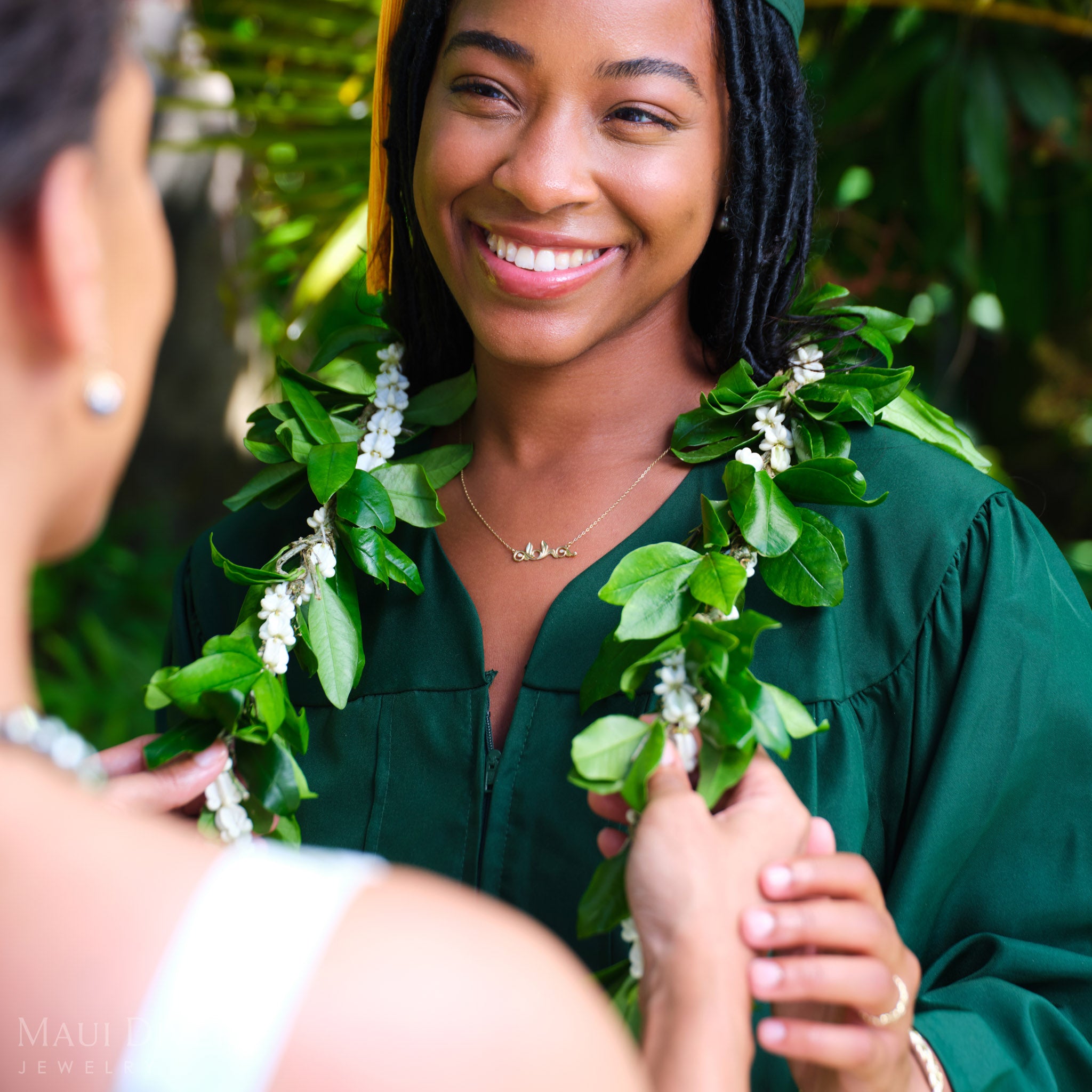 Woman smiling in a green graduation gown wearing a Living Heirloom Aloha necklace in 14k yellow gold with diamond accents, being lei'd by another person with a green lei intertwined with white crown flowers