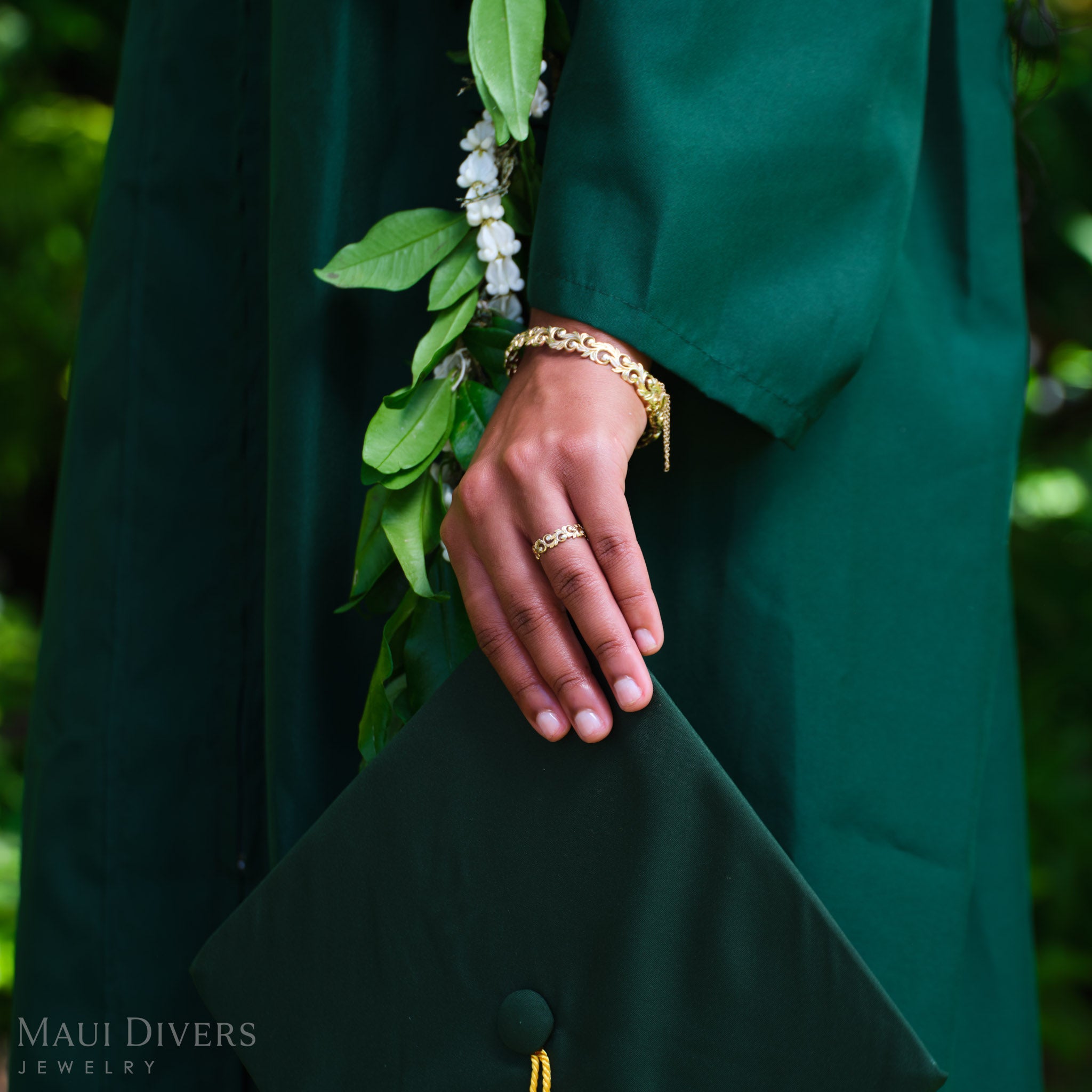 A person in a green graduation gown wearing a Living Heirloom Ring in 14k yellow gold with diamond accents on their ring finger, holding a green graduation cap