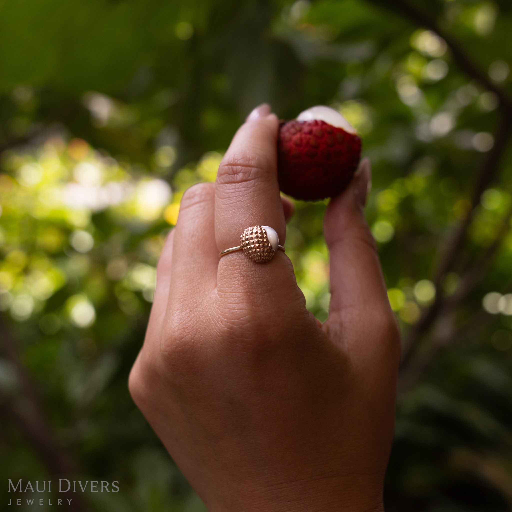 Close-up of a hand wearing a Lychee Mother of Pearl Ring in 14k Two Tone Gold, holding a partially peeled lychee, against a blurred green forest background