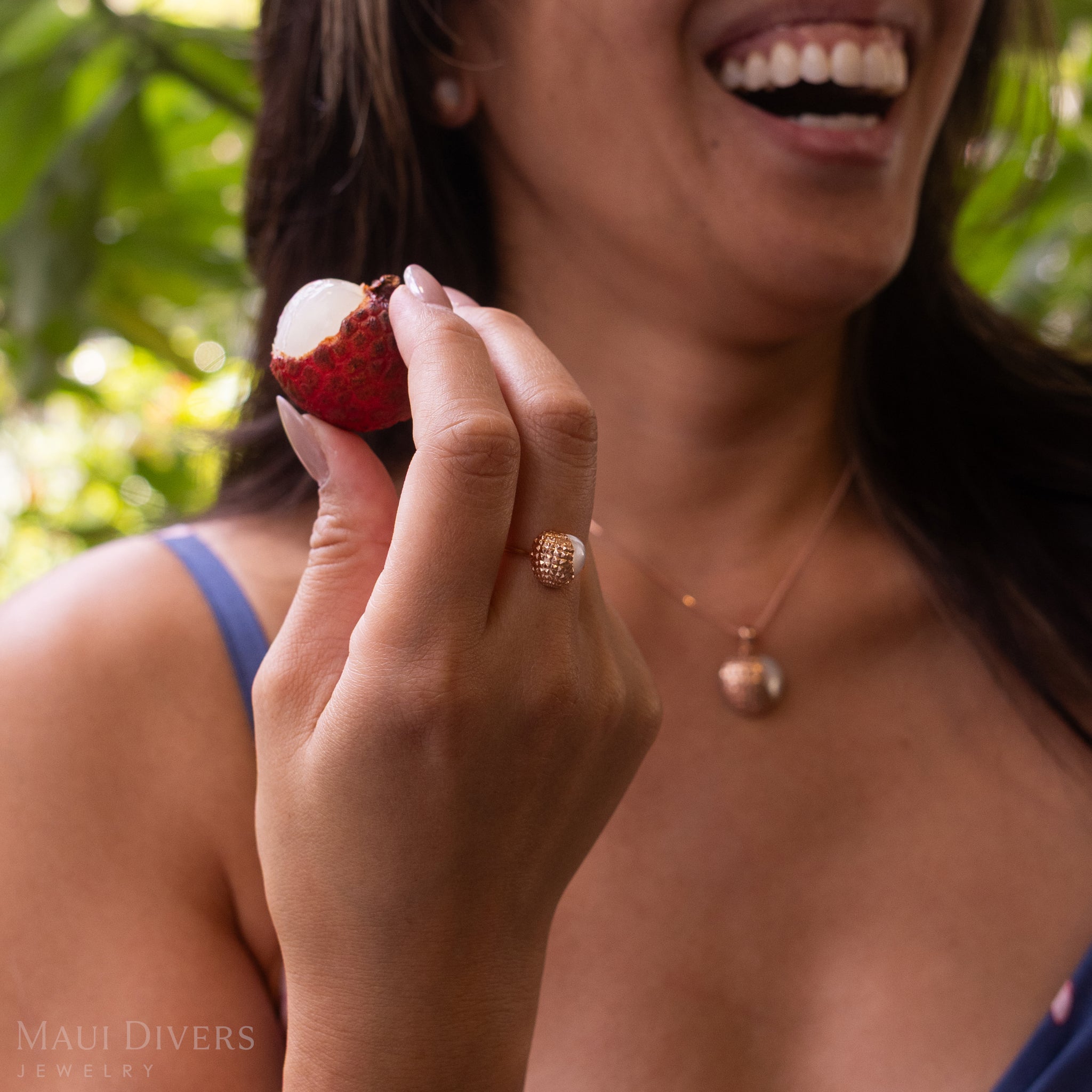 A smiling woman wearing a Lychee Mother of Pearl Ring in 14k Two Tone Gold, holding a partially peeled lychee, against a blurred green forest background