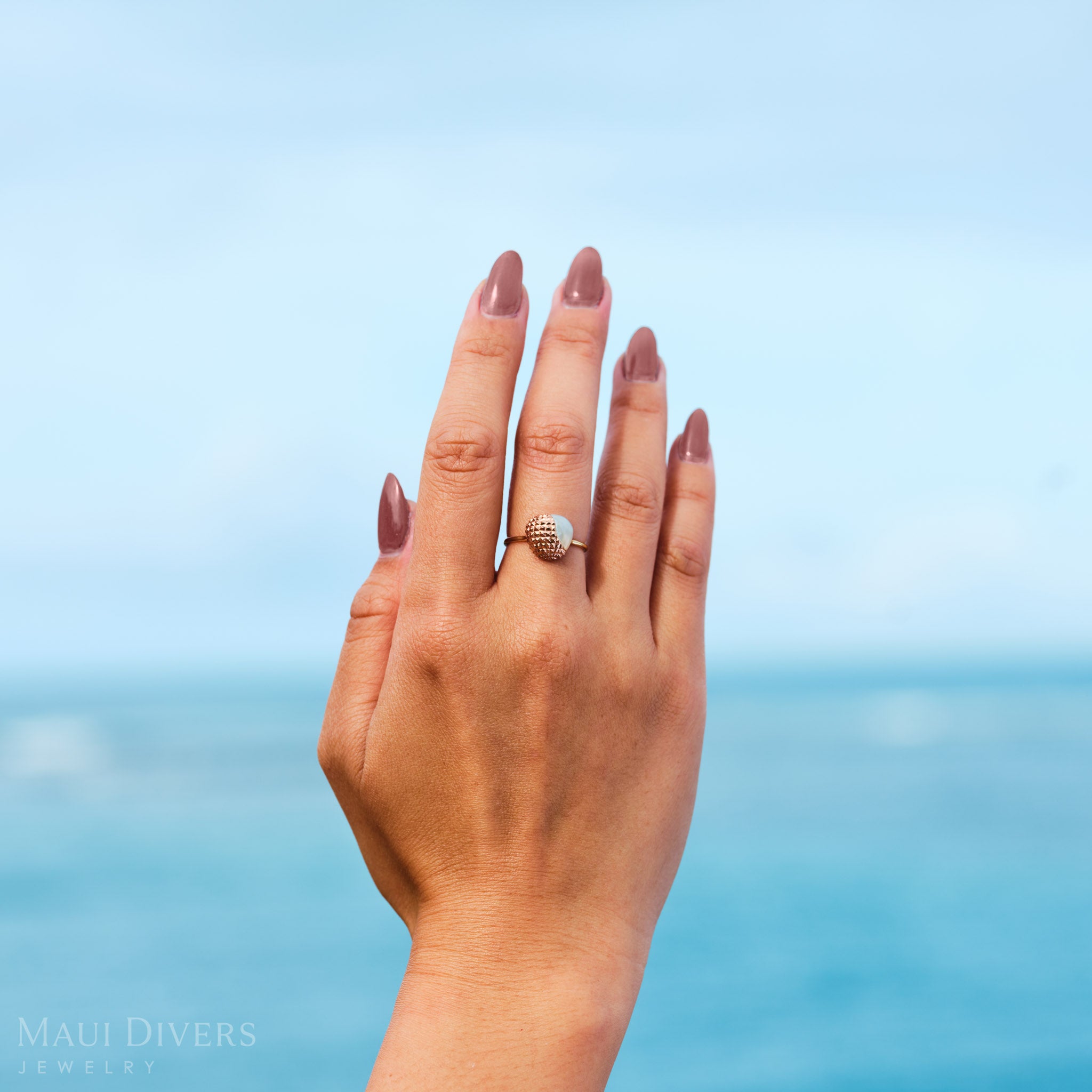 Close-up of a hand wearing a Cute Fruits Lychee Mother of Pearl Ring in two-tone gold, against a blurred ocean background