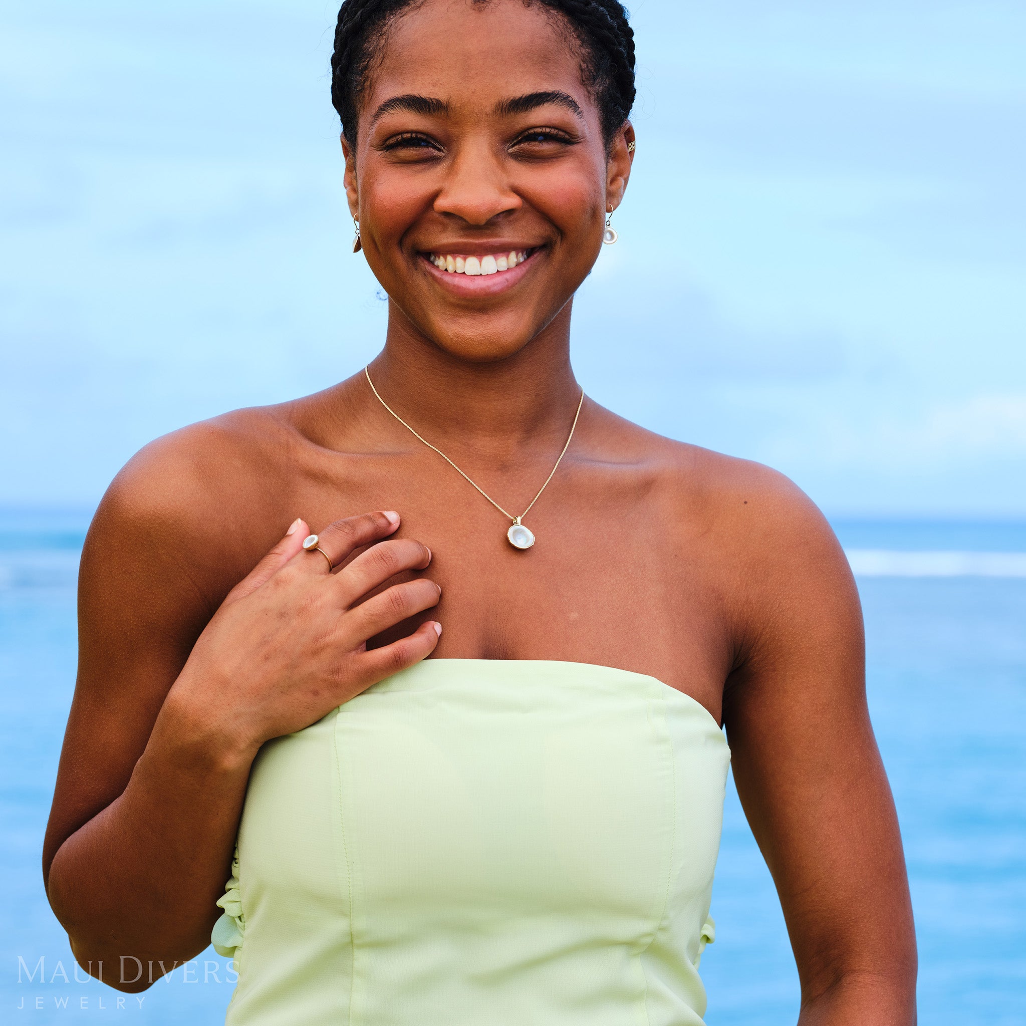 Close-up of a smiling woman in a pastel green dress wearing a Cute Fruits Coconut Mother of Pearl matching set with a pendant, ring, and earrings, all in 14k yellow gold