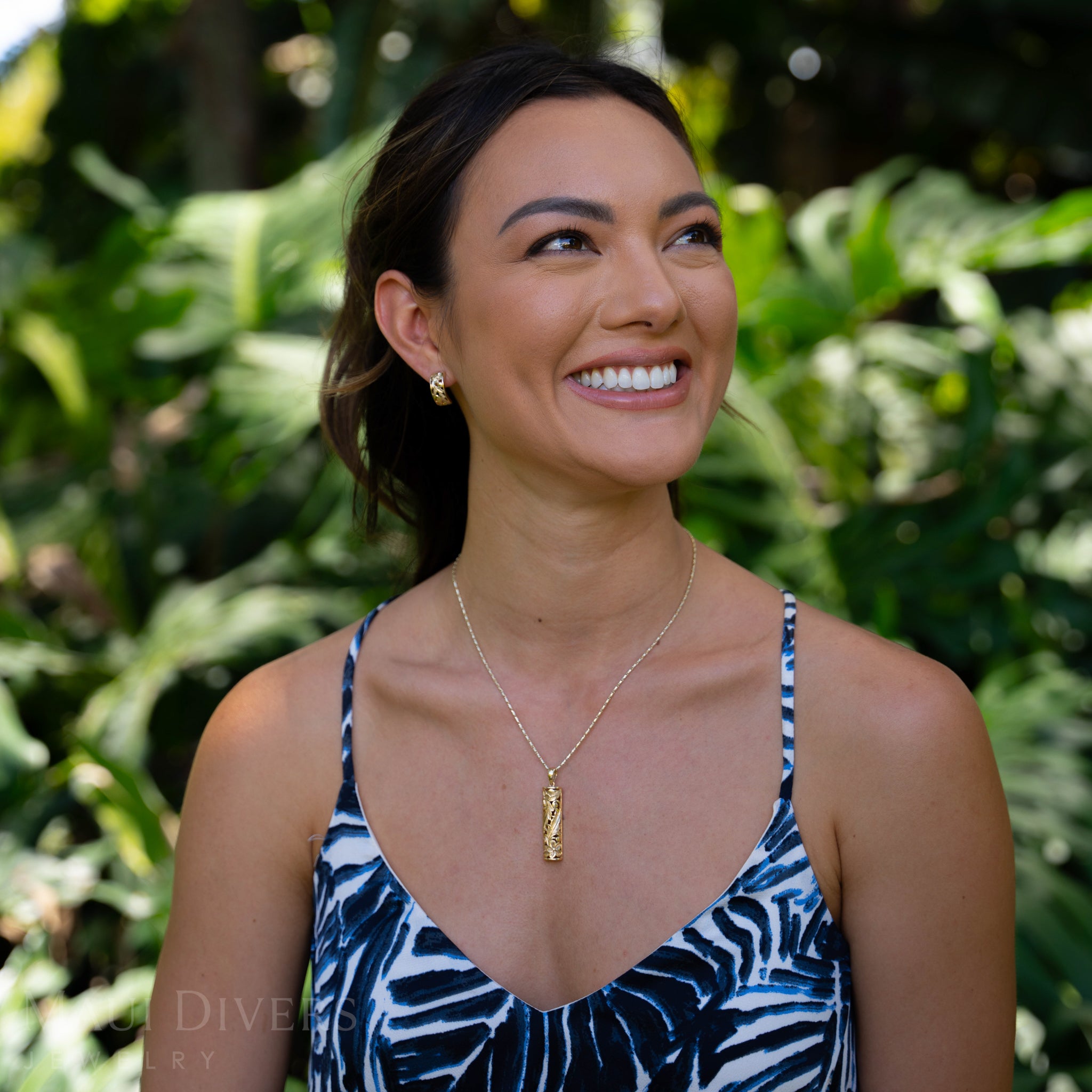 Woman wearing a blue and white patterned top with a gold Hawaiain Heirloom necklace, standing in front of green foliage.