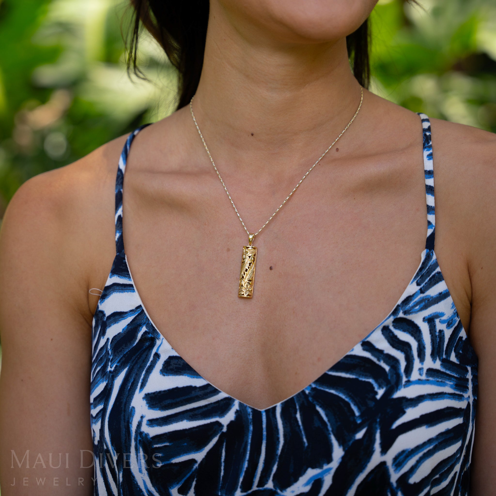 Woman wearing a blue and white patterned top with a gold Hawaiian Heirloom Pendant against a blurred green background