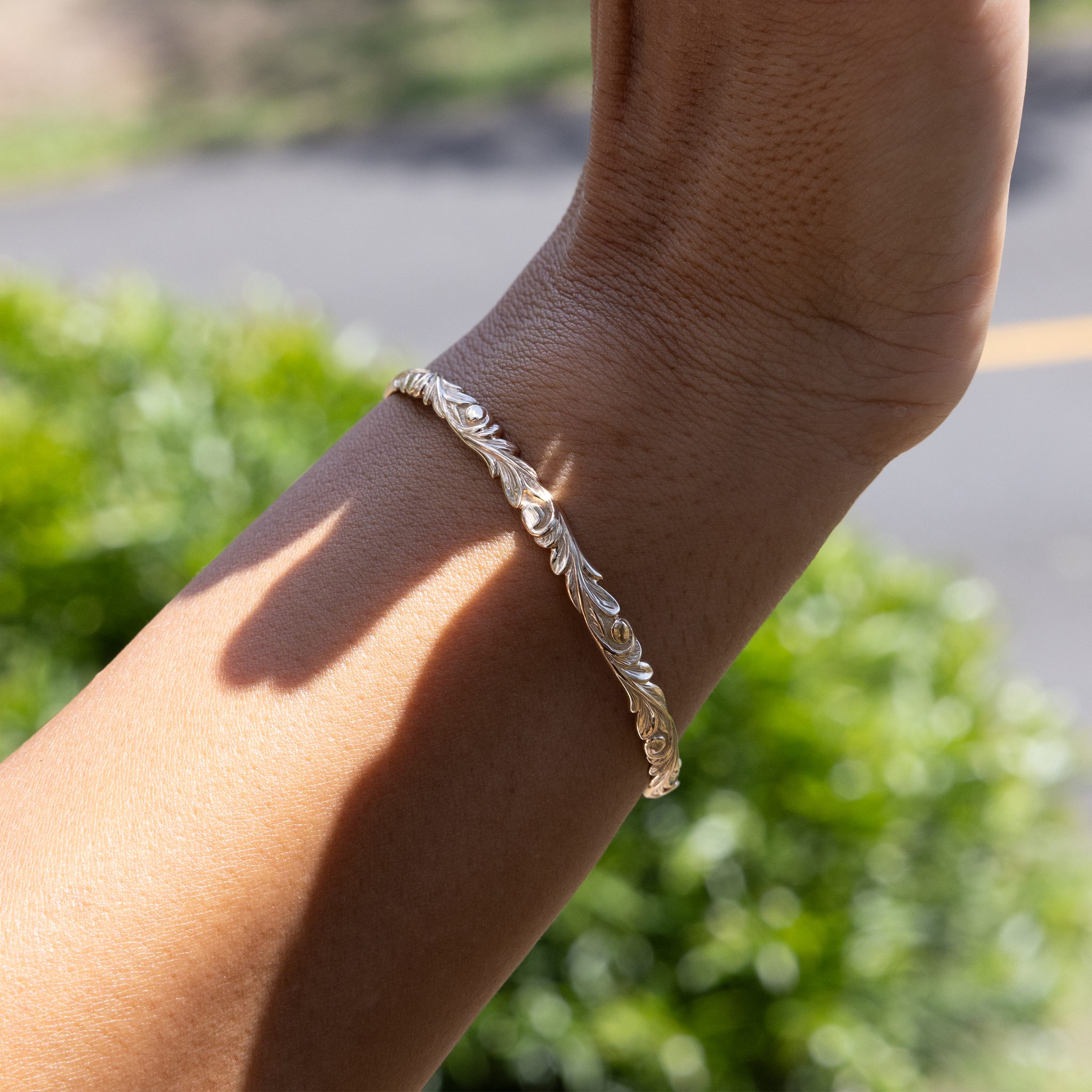 Close up of Hawaiian Heirloom Old English Scroll Bracelet in Gold on a wrist with blurry green background