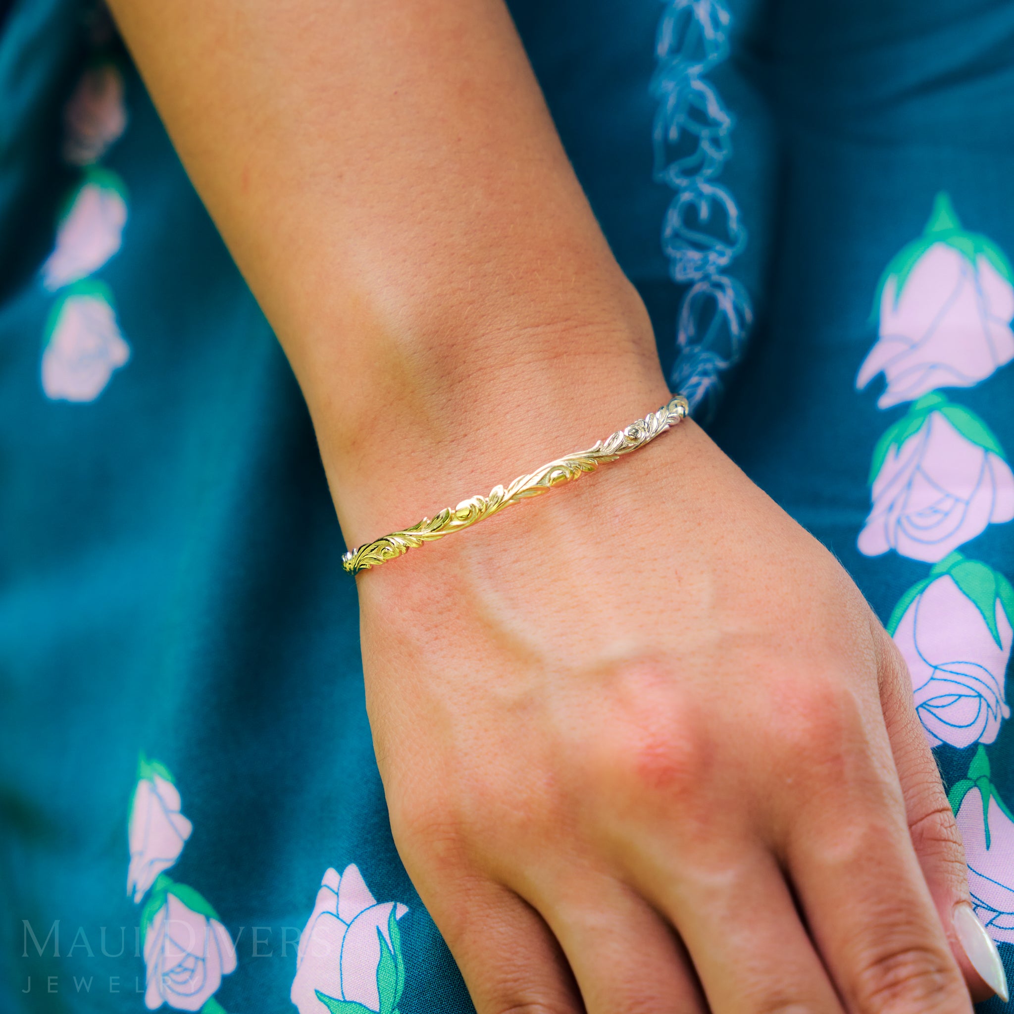 Hawaiian Heirloom Old English Scroll Bracelet in Gold on a wrist with a blue fabric background featuring floral patterns