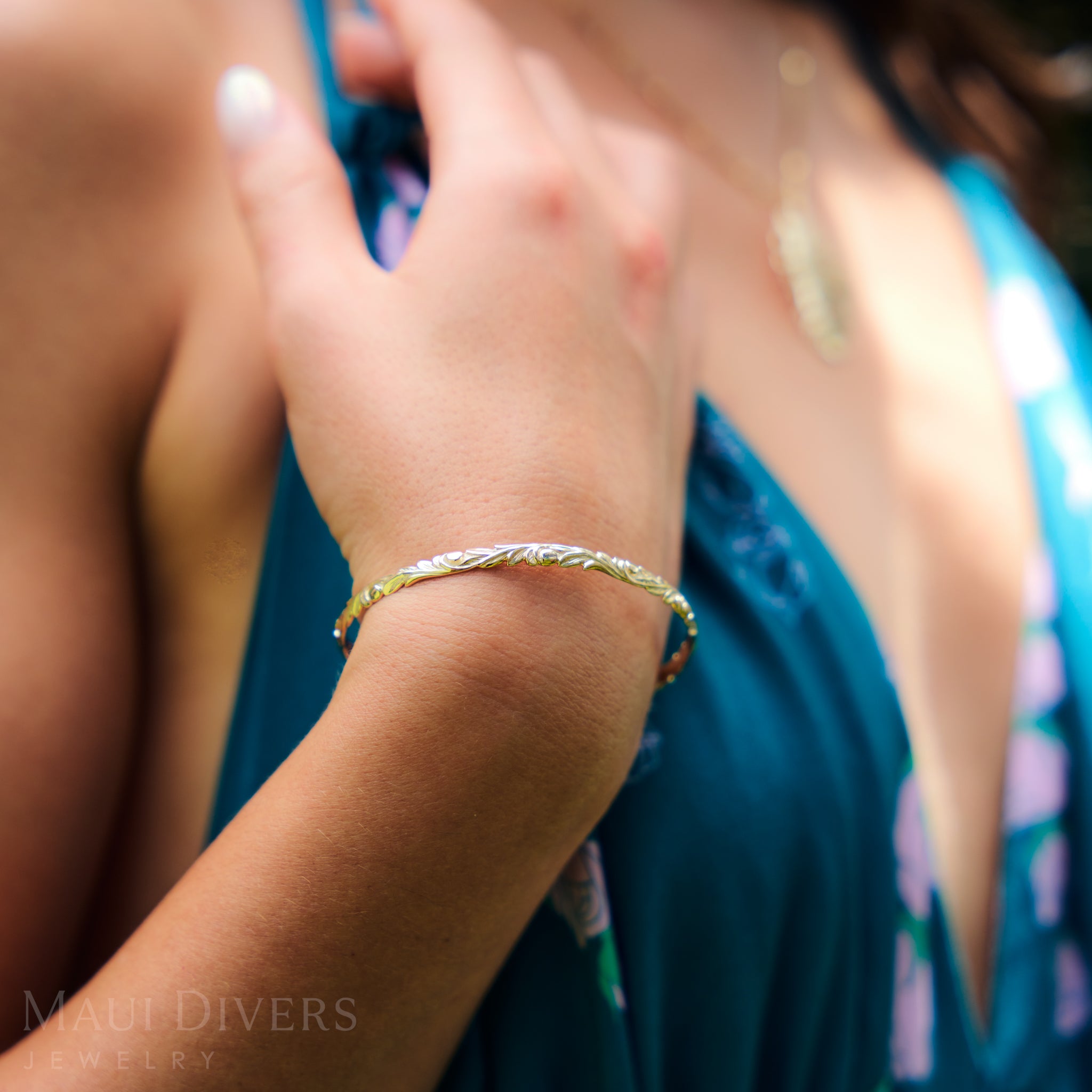 Close-up of a person wearing a Hawaiian Heirloom Old English Scroll Bracelet in Gold with a blurred background