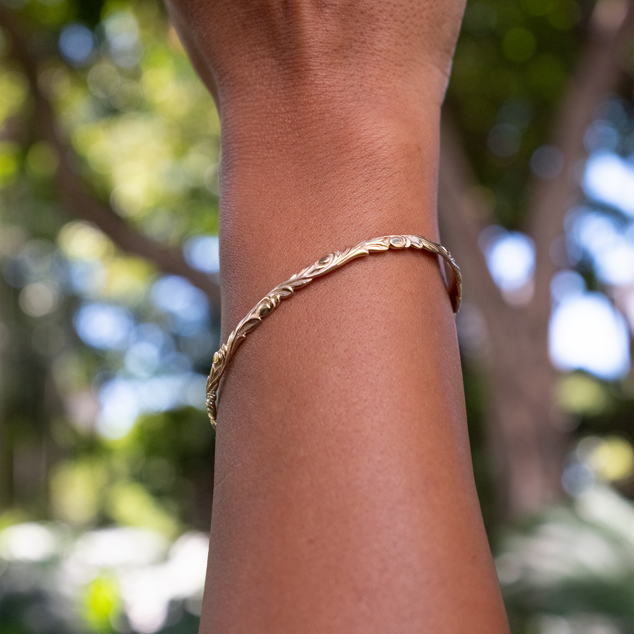 Close up of Hawaiian Heirloom Old English Scroll Bracelet in Gold on a wrist with blurry trees in background
