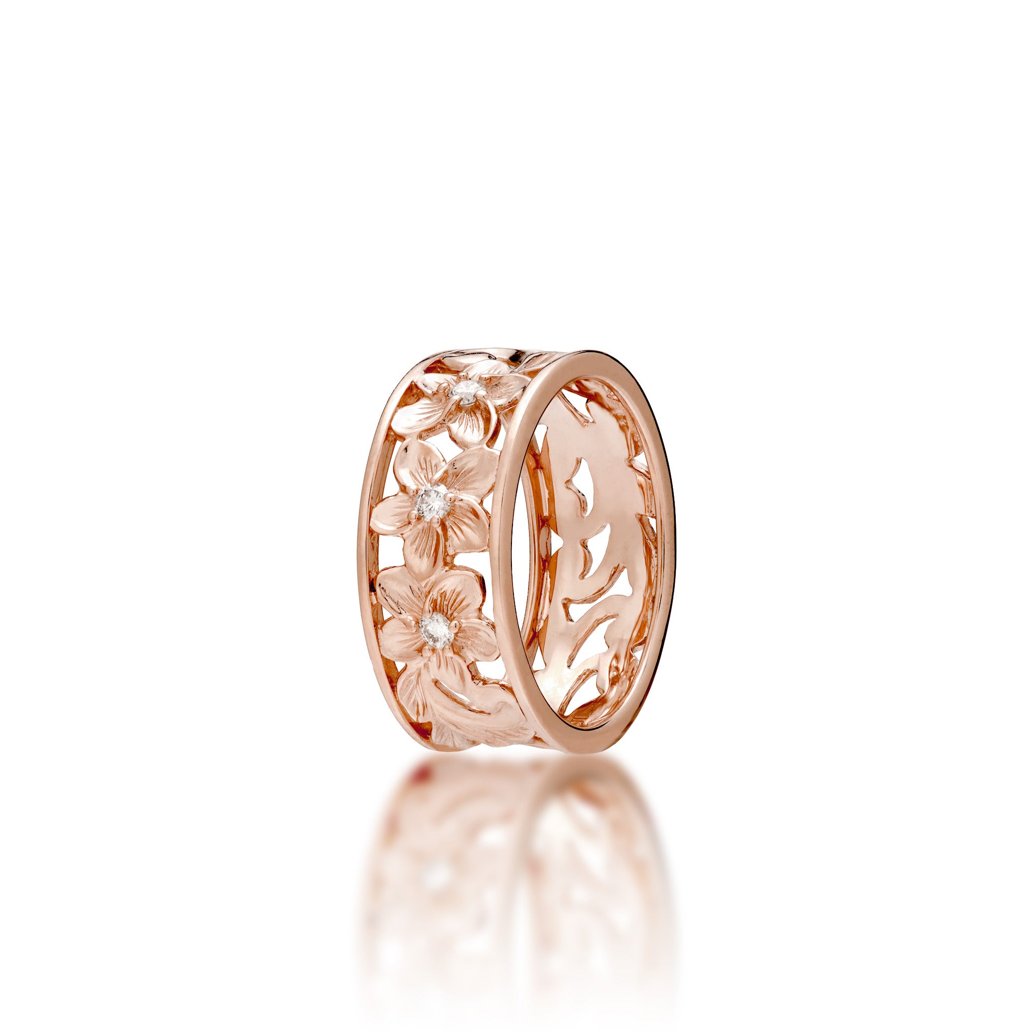 A 14k rose gold Hawaiian Heirloom band ring featuring an maile leaf and fern scrollwork and plumeria flower pattern with a small diamond at the center of each flower, shown on a white background with a soft reflection below.