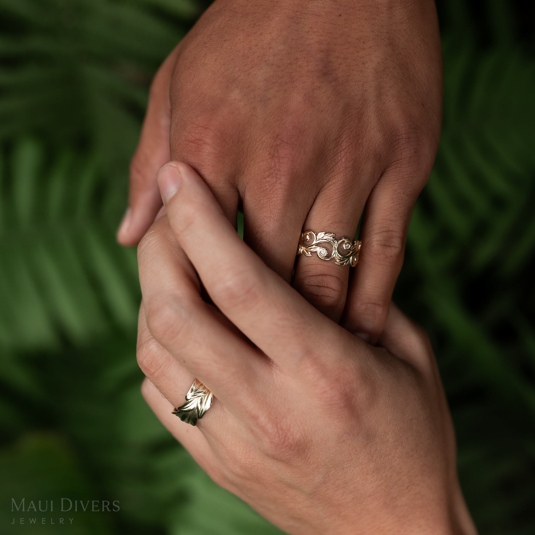Maile and Living Heirloom Rings in Gold with Diamonds on holding hands over blurred leafy background