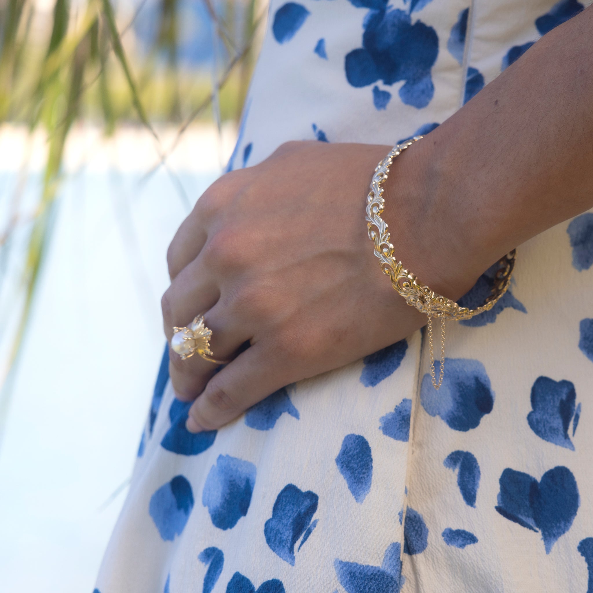 Living Heirloom Hinge Bracelet in 14k Yellow Gold on a wrist with a blurred palm and ocean background