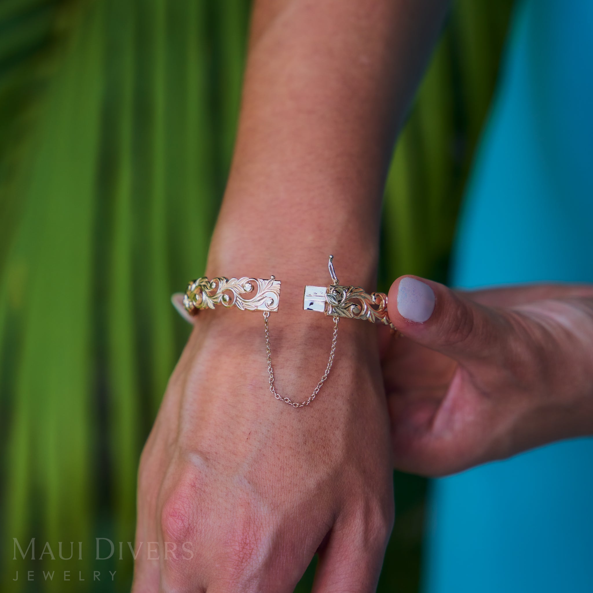 Close-up of a hand wearing a Living Heirloom Hinge Bracelet in 14k Yellow Gold with a blurred green and blue background.