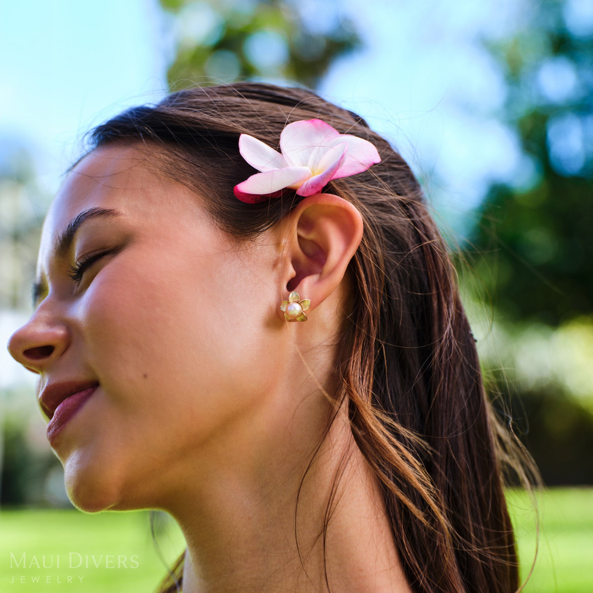 Close-up of a smiling woman wearing a Pick A Pearl Plumeria Earring in 14k yellow gold with a pink pearl on her left ear, against a blurred park background