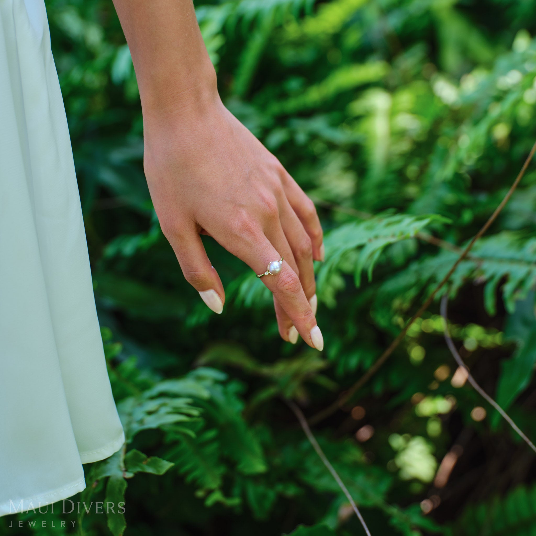 Woman wearing a Pick A Pearl Crown Ring in 14k yellow gold with diamonds and a white pearl, against a blurred leafy background