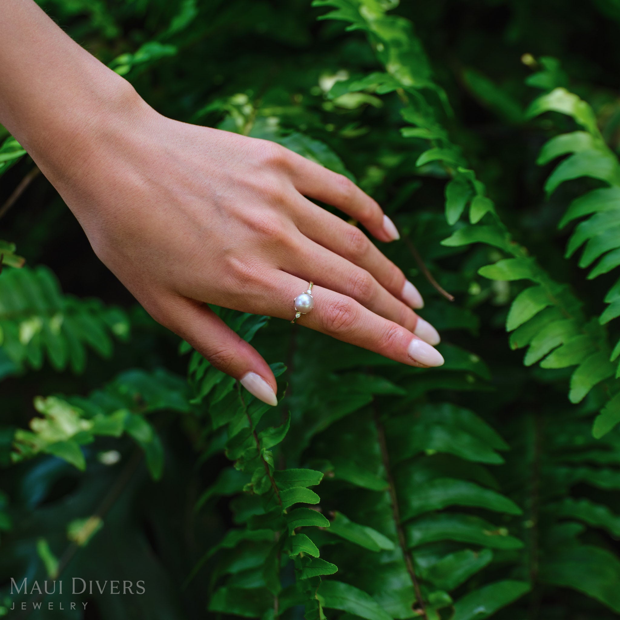 Close-up of a Pick A Pearl Crown Ring in 14k yellow gold with diamonds and a white pearl, worn on a hand, against a leafy background