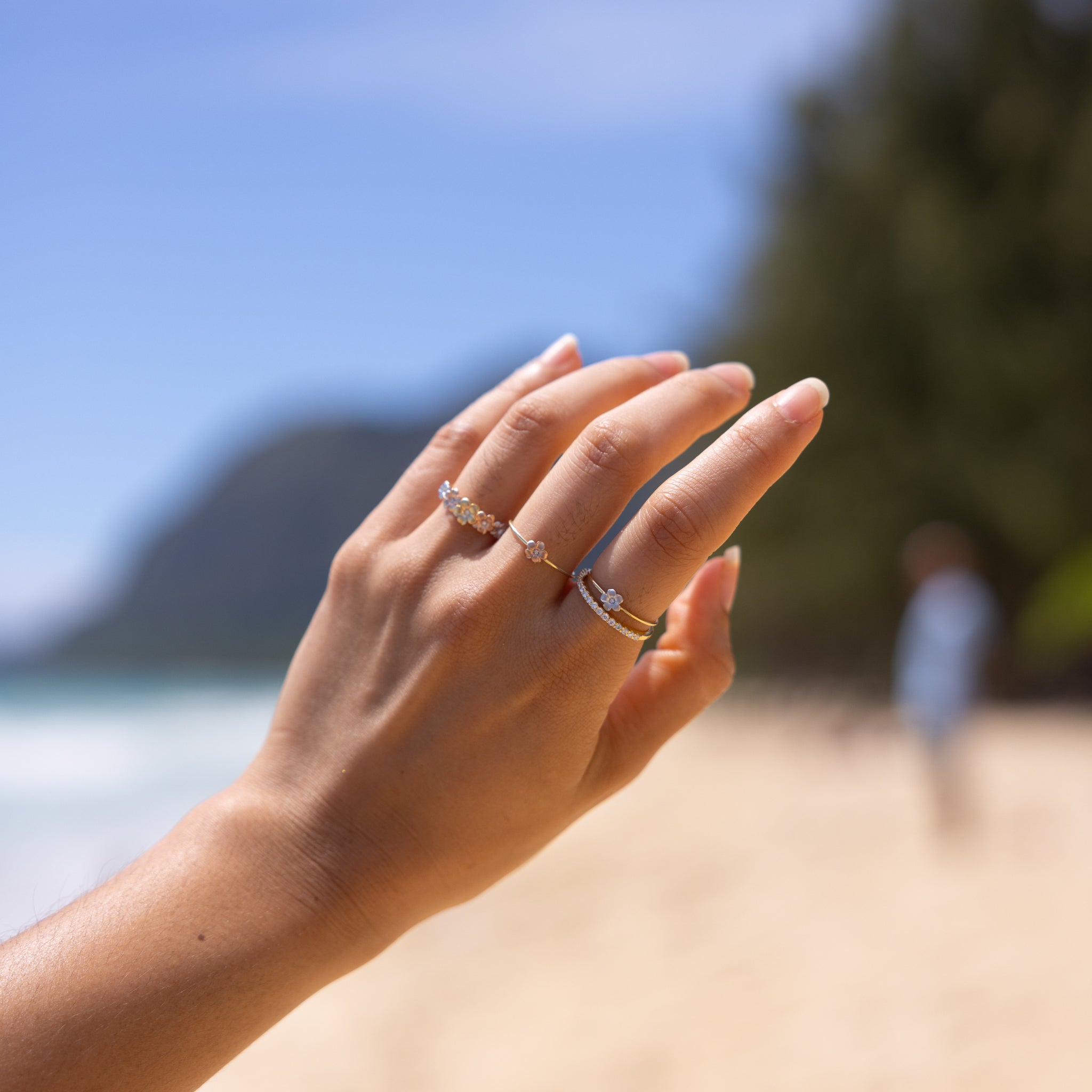 Hand wearing multiple Plumeria Rings in Gold with Diamonds against a blurred ocean and mountain background