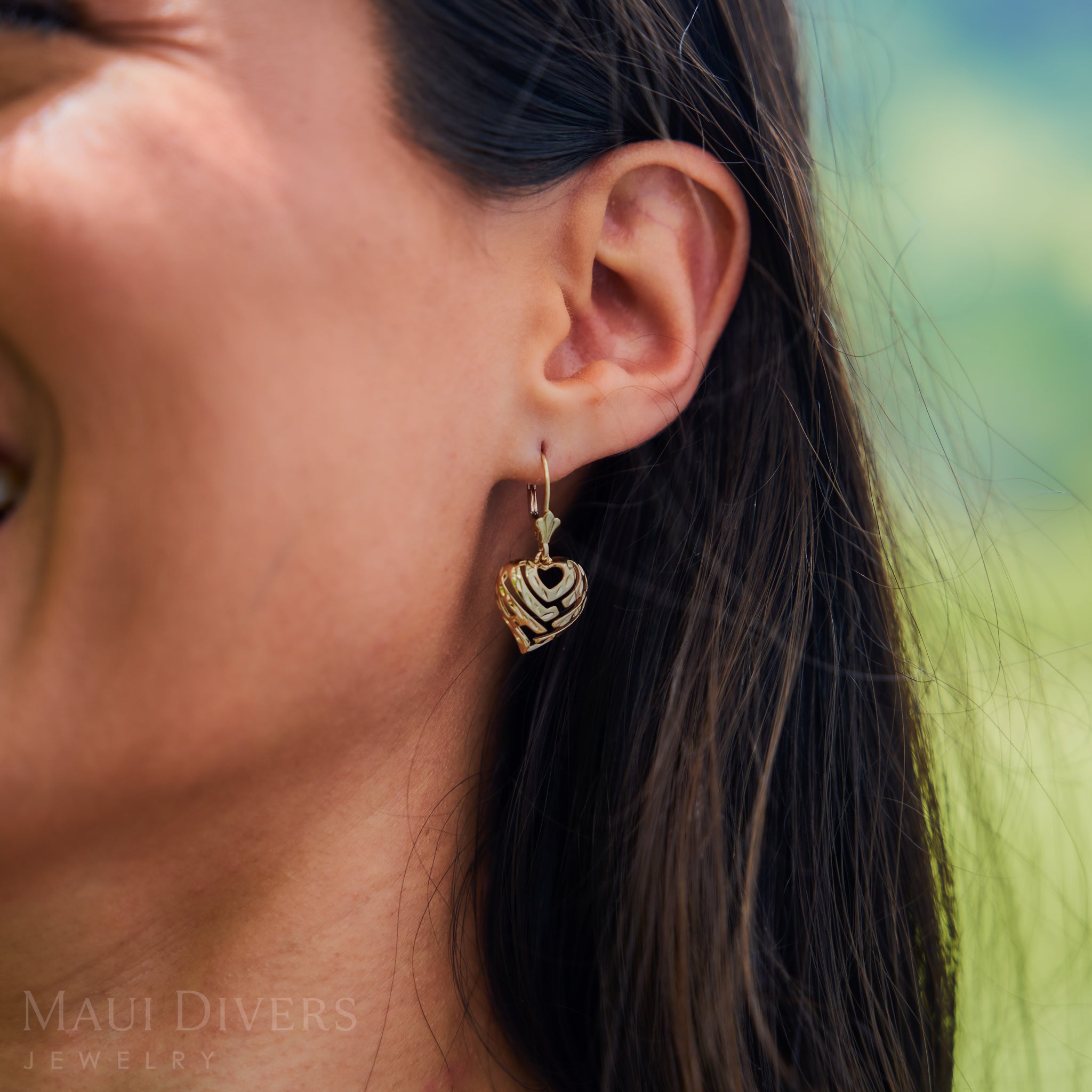 Close-up of a person wearing Aloha Heart Earrings in 14k Yellow Gold with a blurred natural background