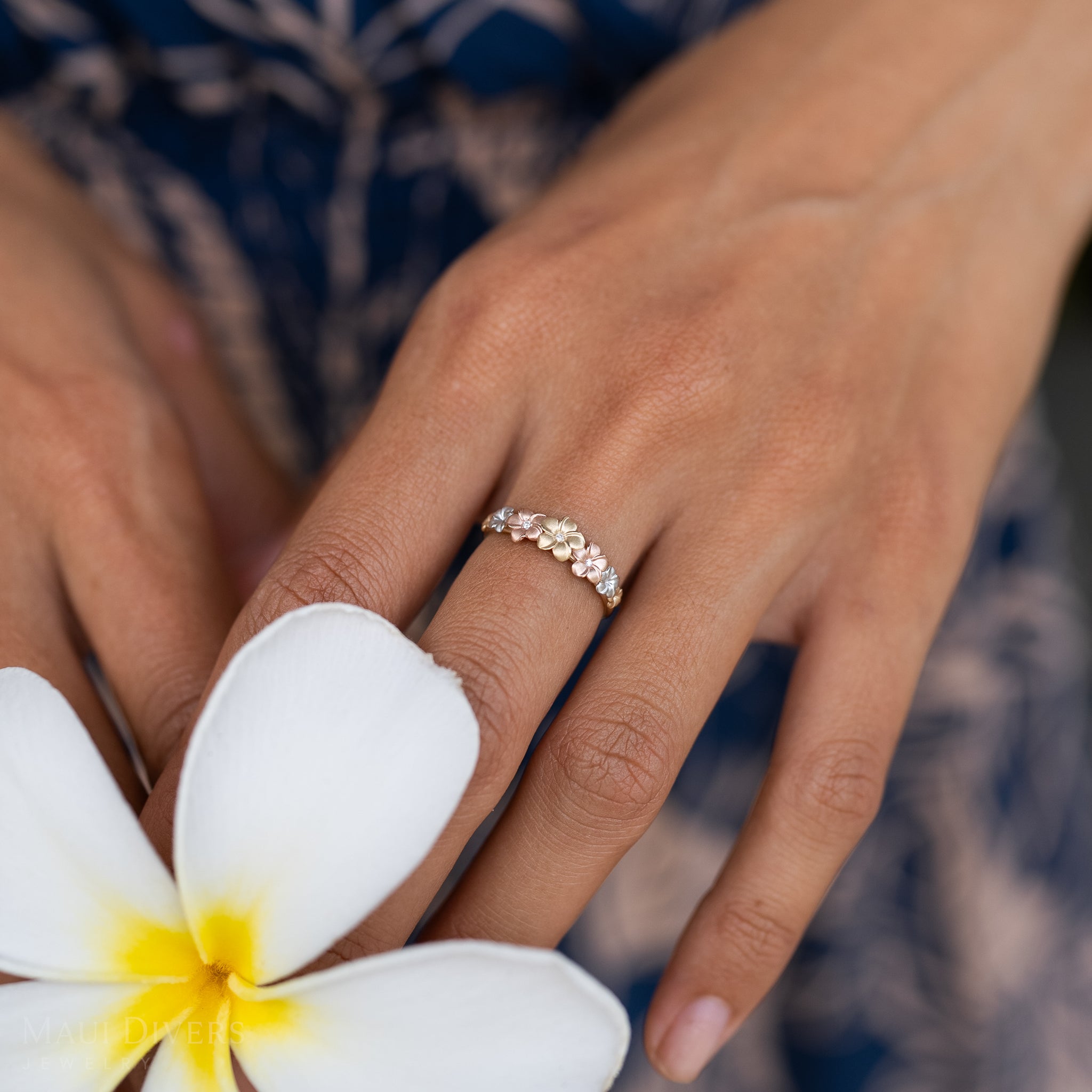 Close up of hand wearing a 6mm 7-Plumeria Ring in Tri Color Gold with Diamonds holding a white plumeria