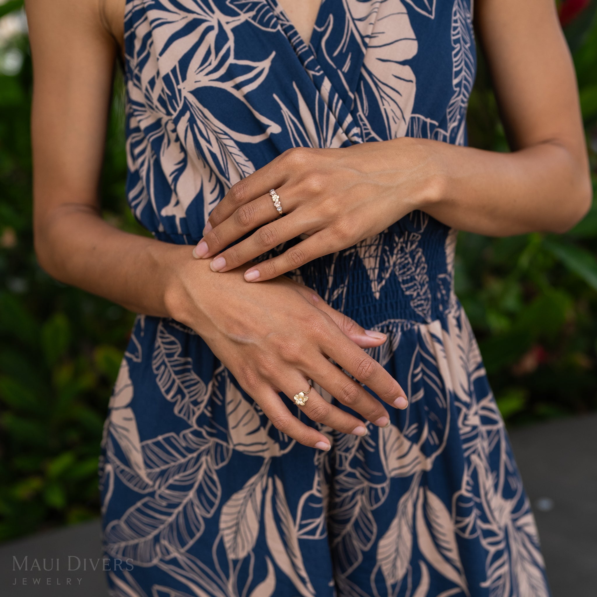 Hands wearing Plumeria Rings in Tri Color Gold with Diamonds against blue and beige pattern jumpsuit and greenery