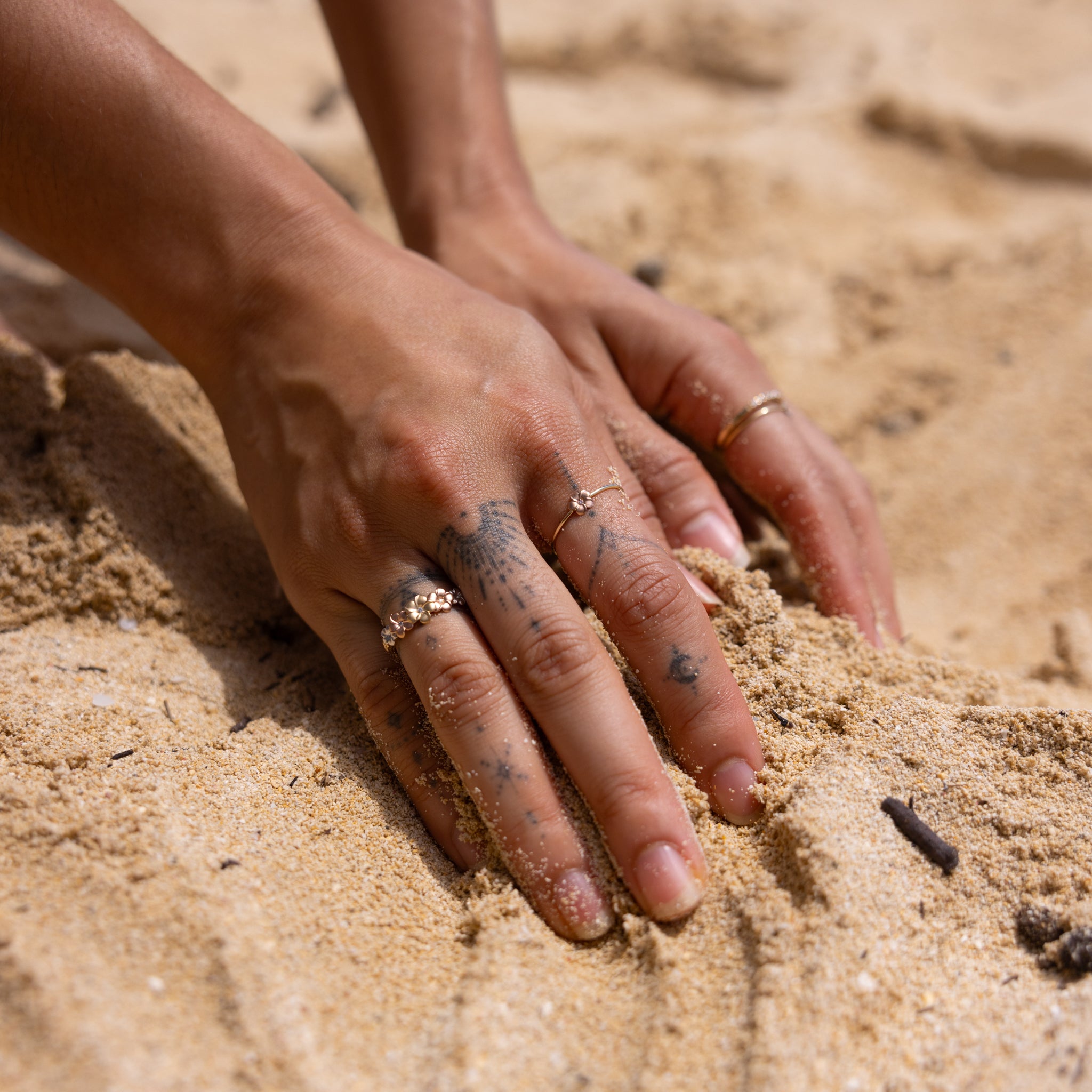 Hands in the sand wearing multiple Plumeria Rings in Gold with Diamonds