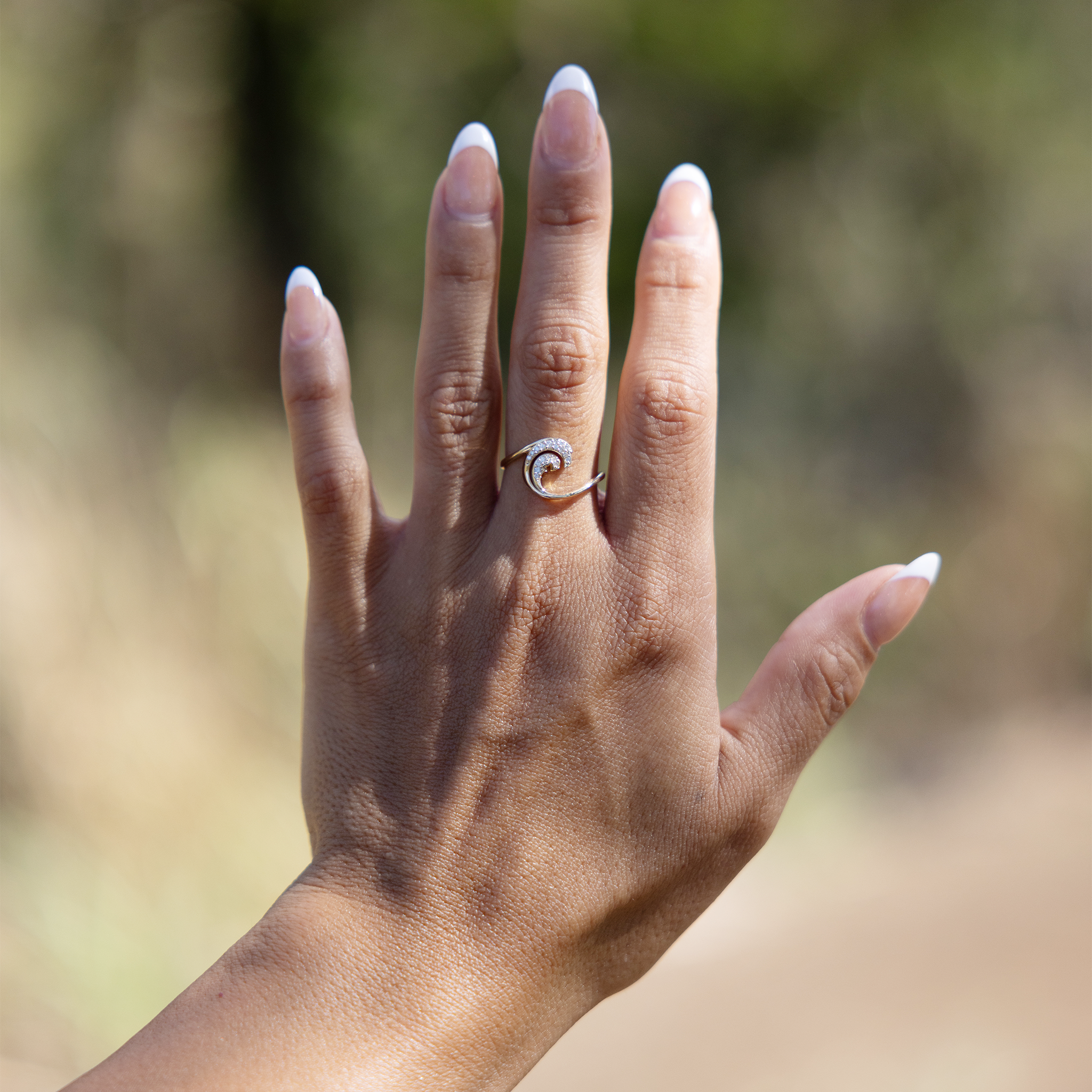 Hand up against a blurred nature background wearing a 12mm Nalu Ring in Gold with Diamonds