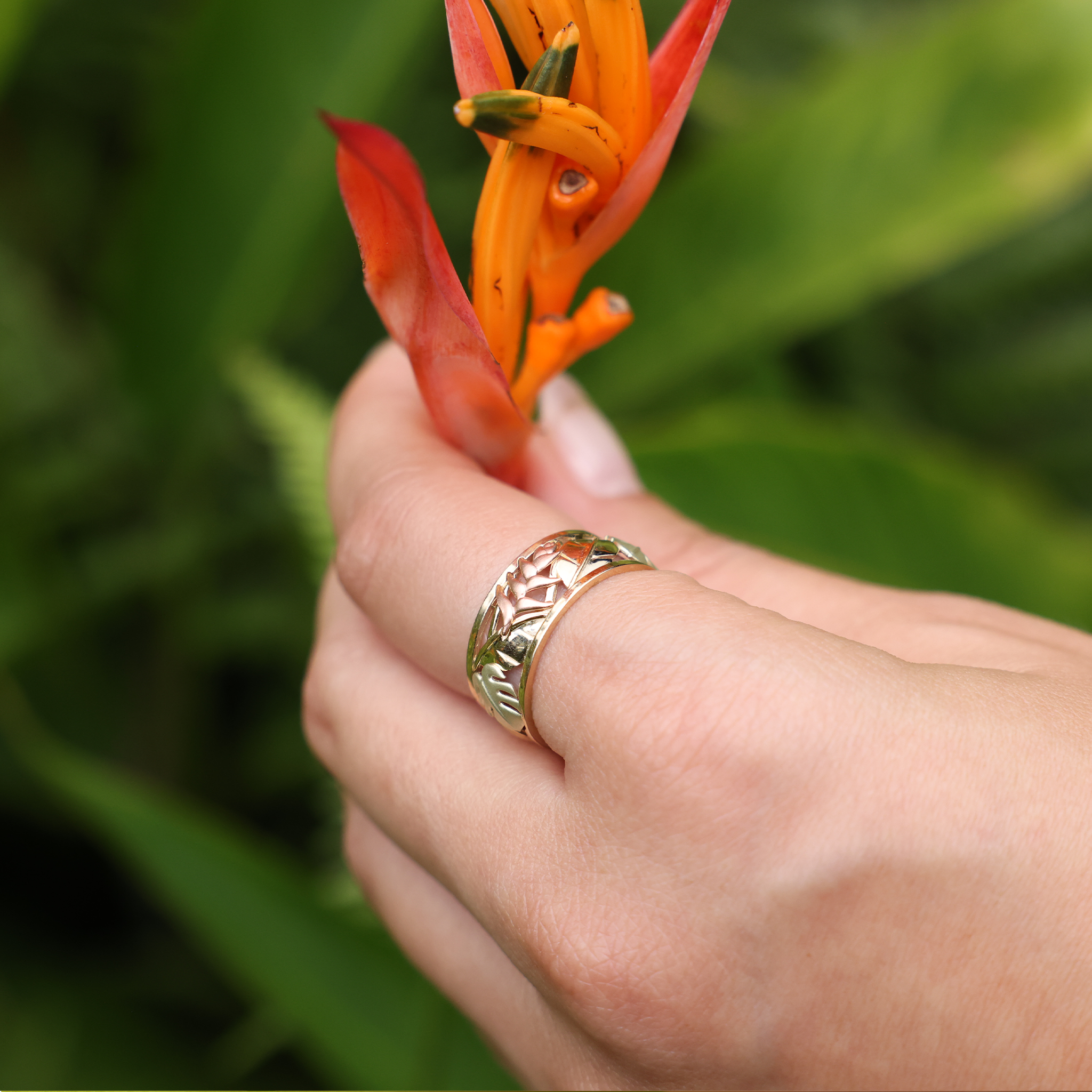 A womanʻs hand with a Hawaiian Gardens Monstera & Heliconia Ring in Tri Color Gold - 8mm holding a flower- Maui Divers Jewelry