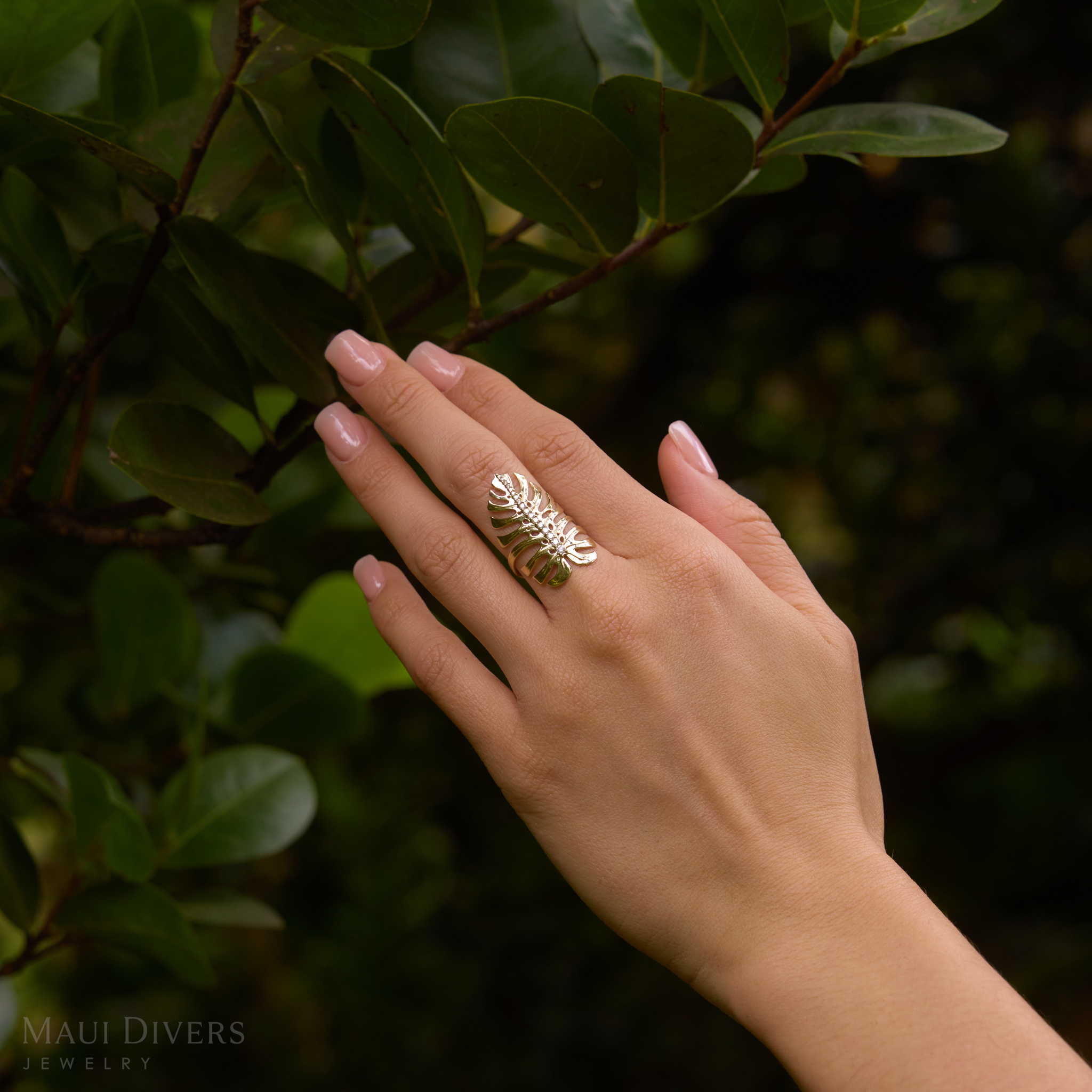 32mm Monstera Ring in Gold with Diamonds on hand up against dark green leaves