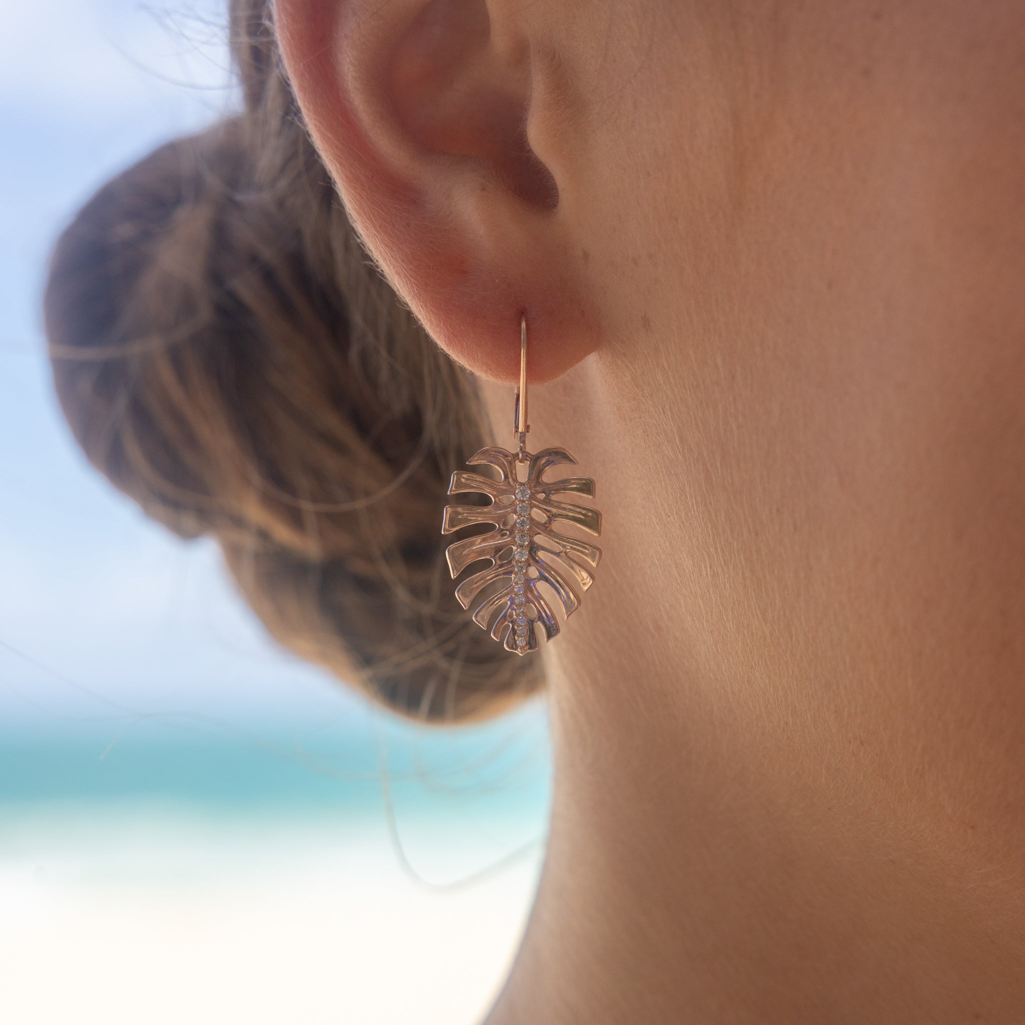 Close-up of a person wearing Monstera Earrings in Rose Gold with Diamonds with a blurred beach background