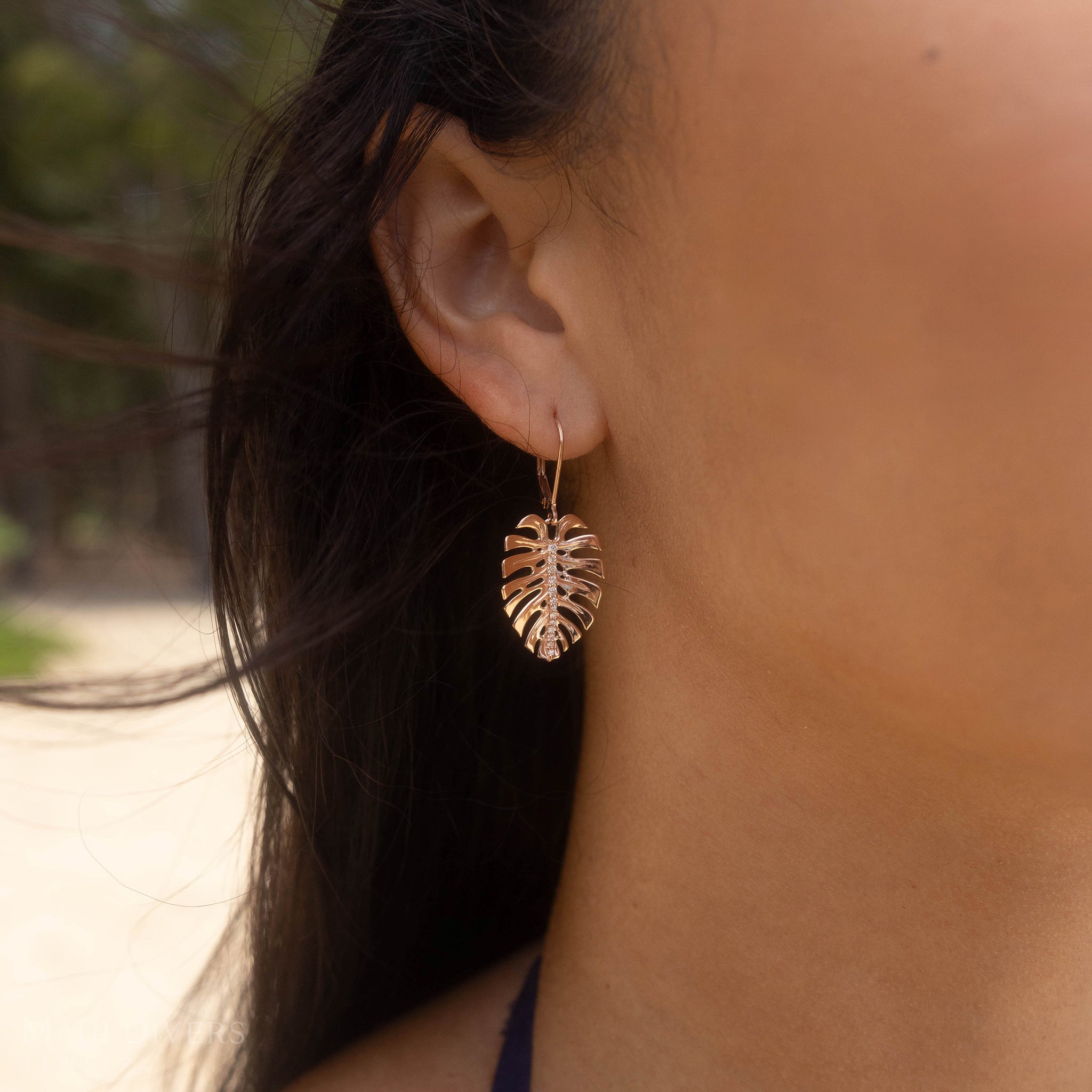 Close up of 23mm Monstera Earrings in Rose Gold with Diamonds on ear with blurred beach in the background
