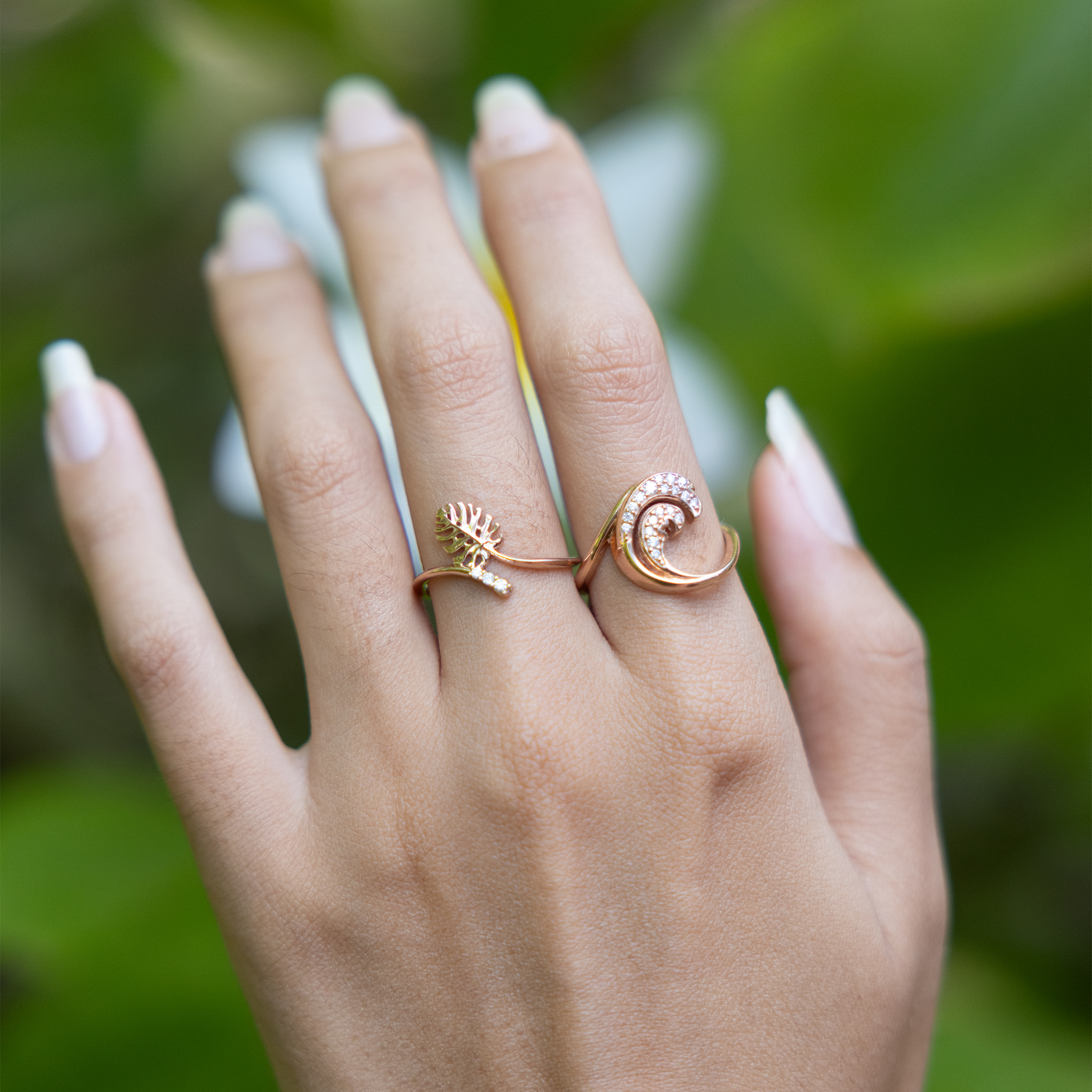 Close up of 9mm Monstera and Nalu Rings in Rose Gold with Diamonds on a hand over blurred leaves