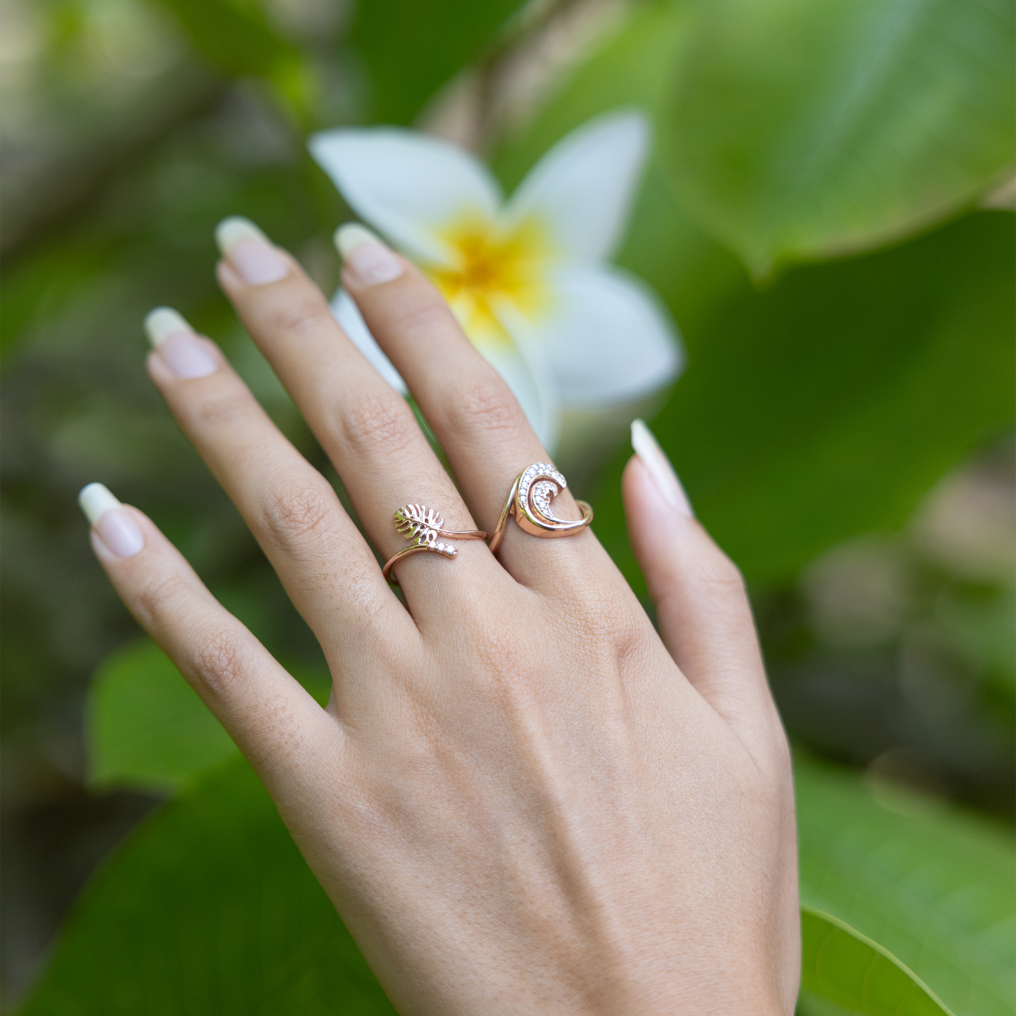 Close up of 9mm Monstera Ring in Rose Gold with Diamonds on a hand over blurred plumeria flower and leaves