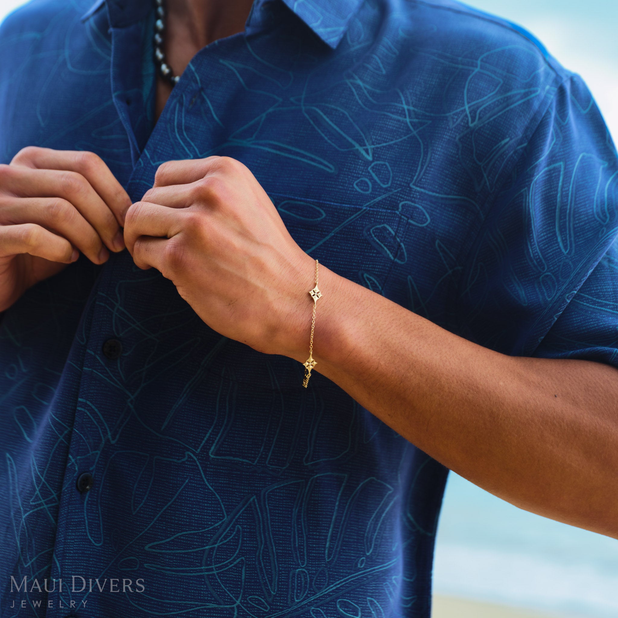 Person wearing a blue patterned shirt with a gold Hawaiian quilted star bracelet, close-up of hands buttoning shirt.