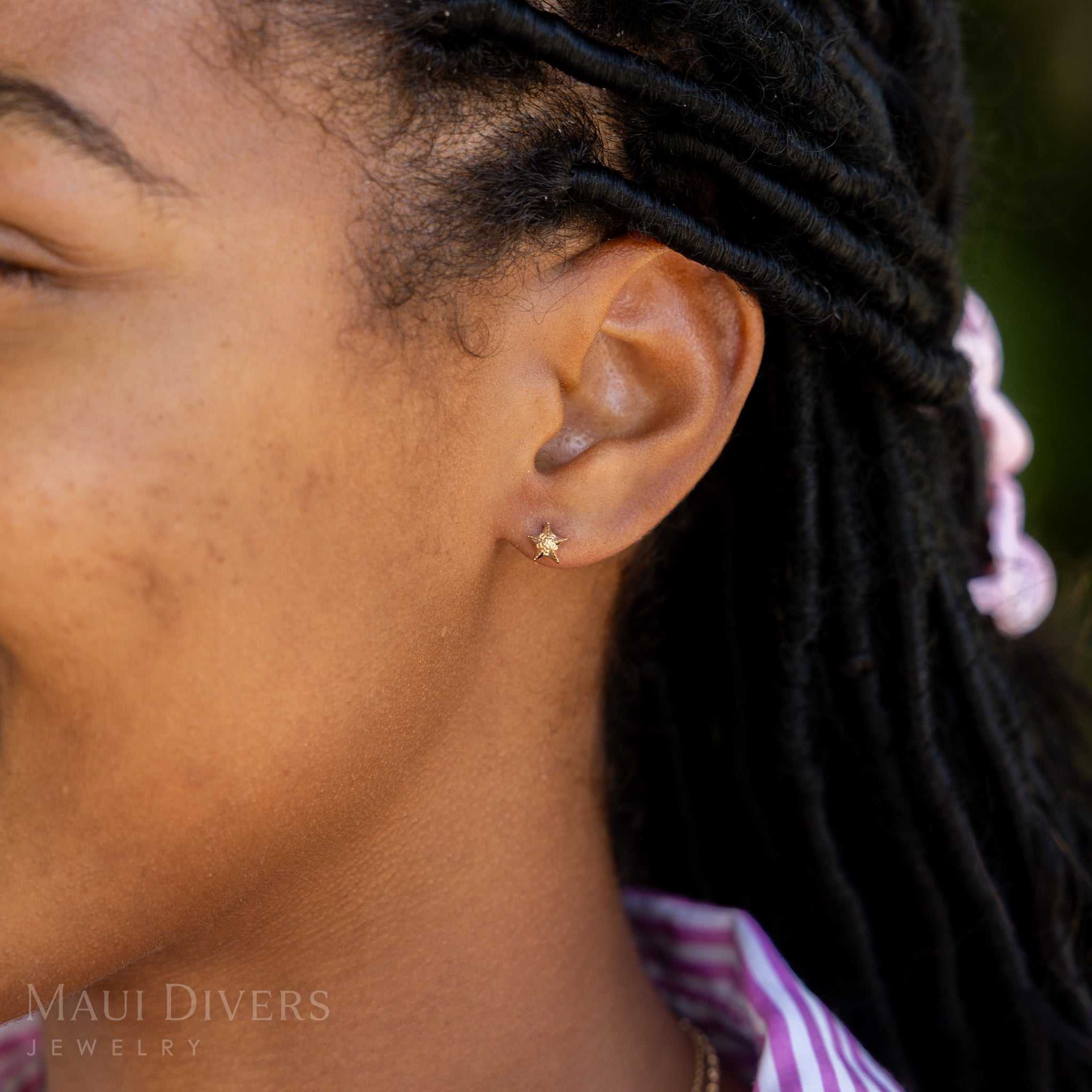 A side view of a smiling woman in a pink and white stripe top wearing a Starfish Earring in 14k yellow gold, against a blurred green background
