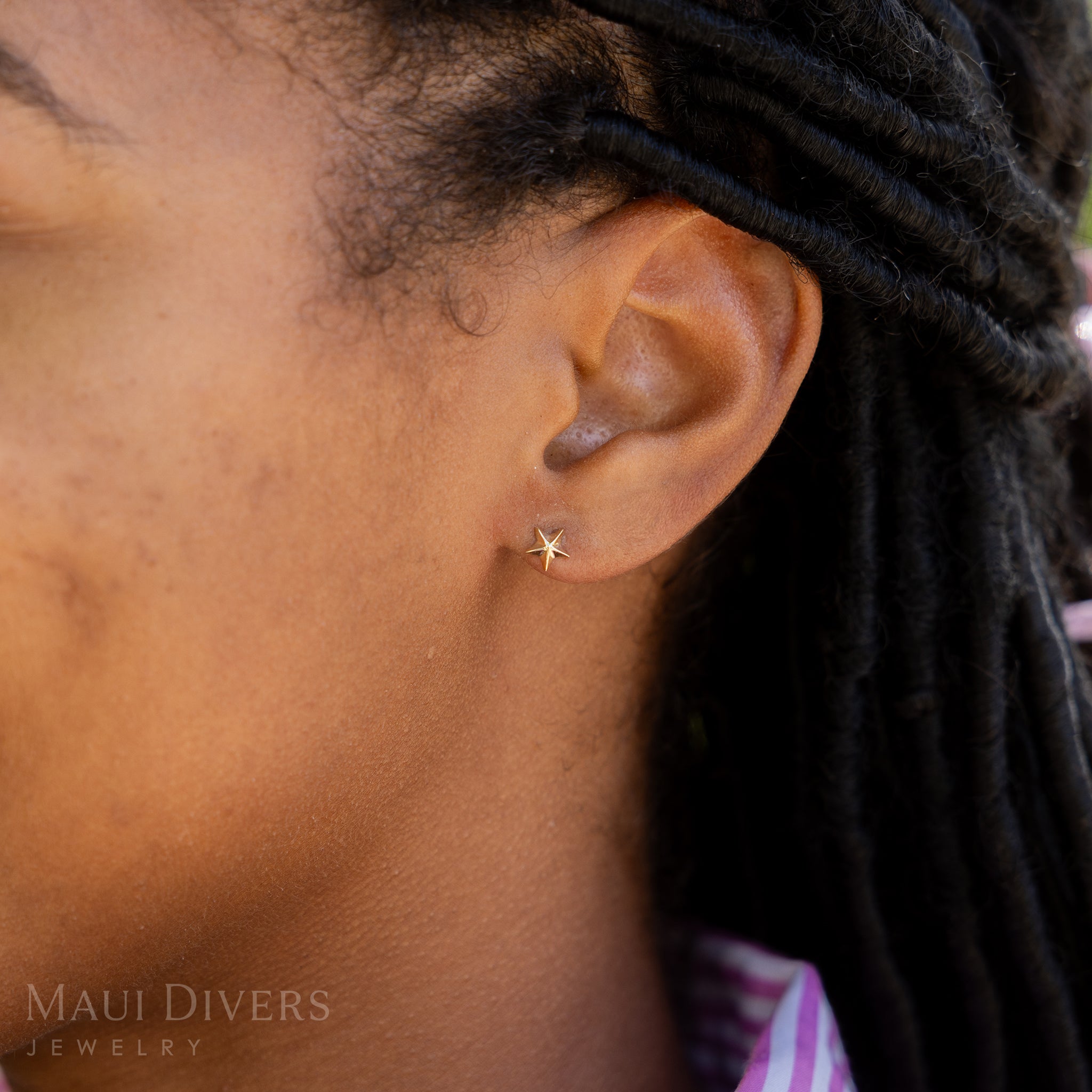 A side view close-up of a woman wearing a Starfish Earring in 14k yellow gold, against a blurred leafy green background
