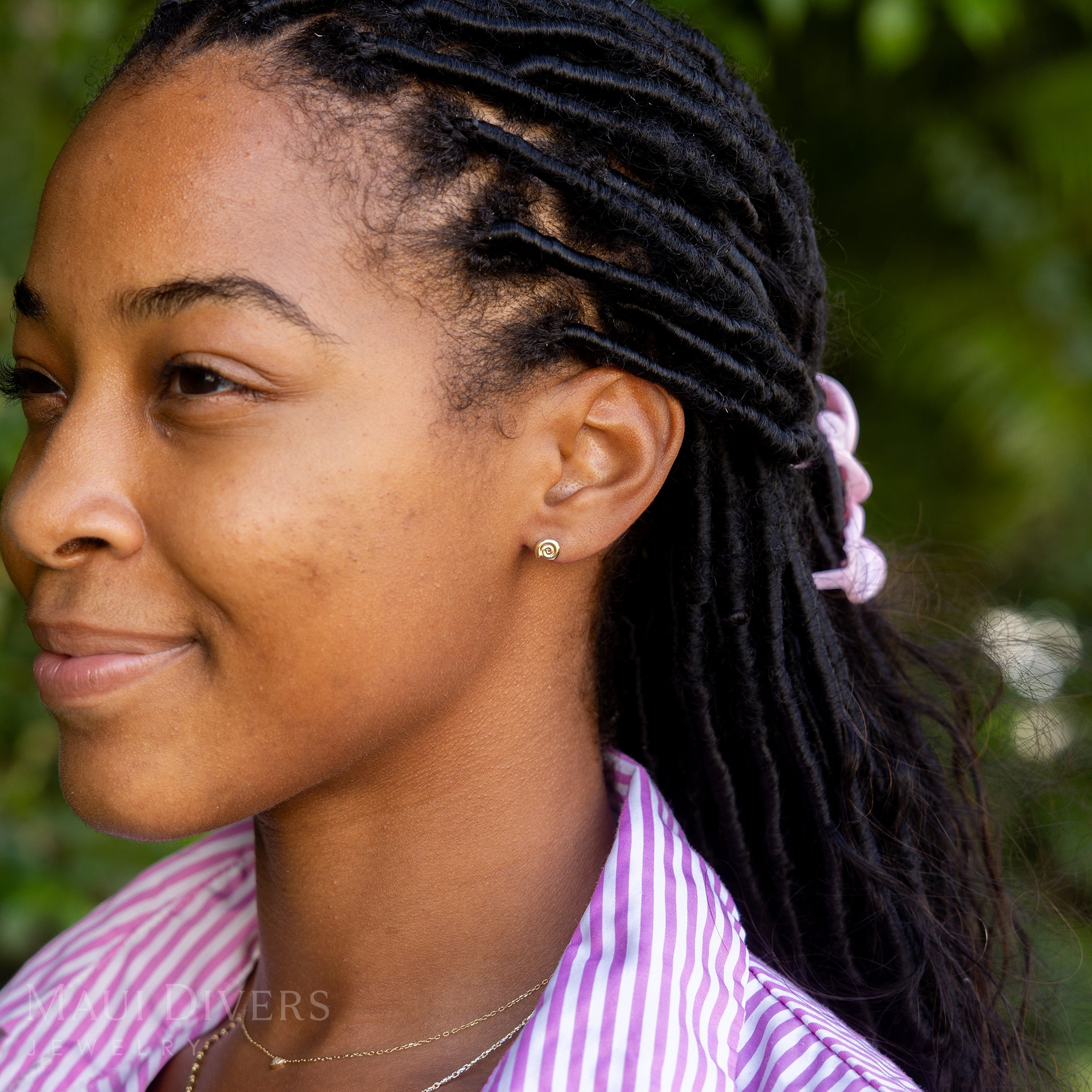 A side view of a smiling woman in a pink and white stripe top wearing a Conch Shell Earring in 14k yellow gold, paired with a tiny diamond necklace, against a blurred leafy green background