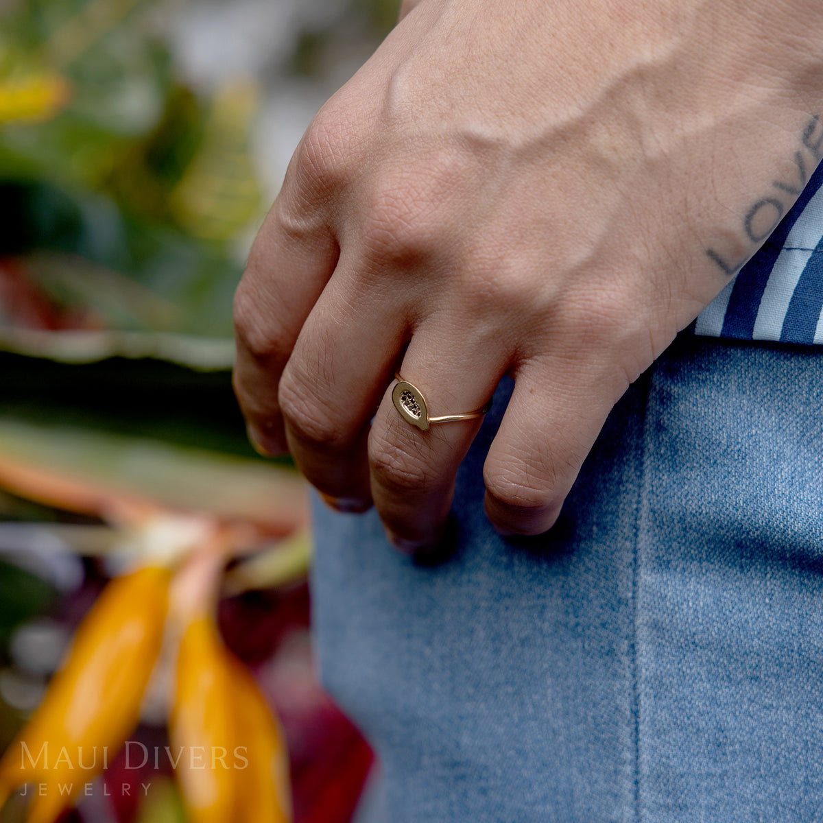 A close-up of a person with their hand against their leg wearing a Cute Fruits Papaya Pendant in Ring in 14k yellow gold with black diamonds on their hand, against a blurred tropical floral background
