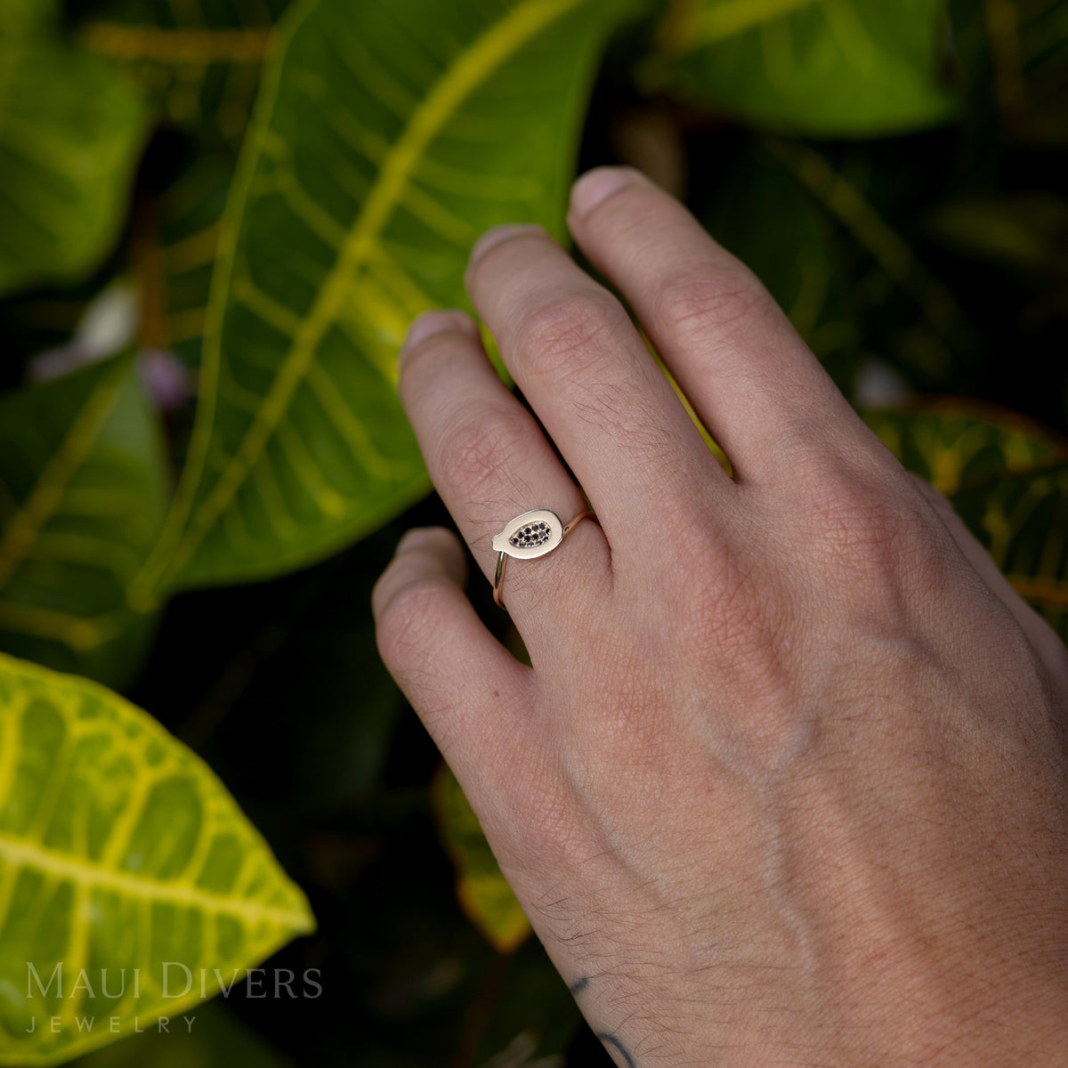 A close-up of a person with their hand against their leg wearing a Cute Fruits Papaya Pendant in Ring in 14k yellow gold with black diamonds on their hand, against a blurred leafy green background