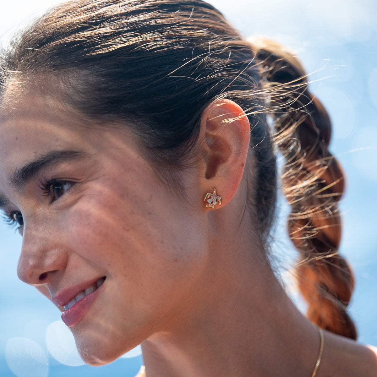 Close-up of a smiling woman wearing a gold sea turtle earring with plumerias in rose and white gold on its back, against a blue skybackground