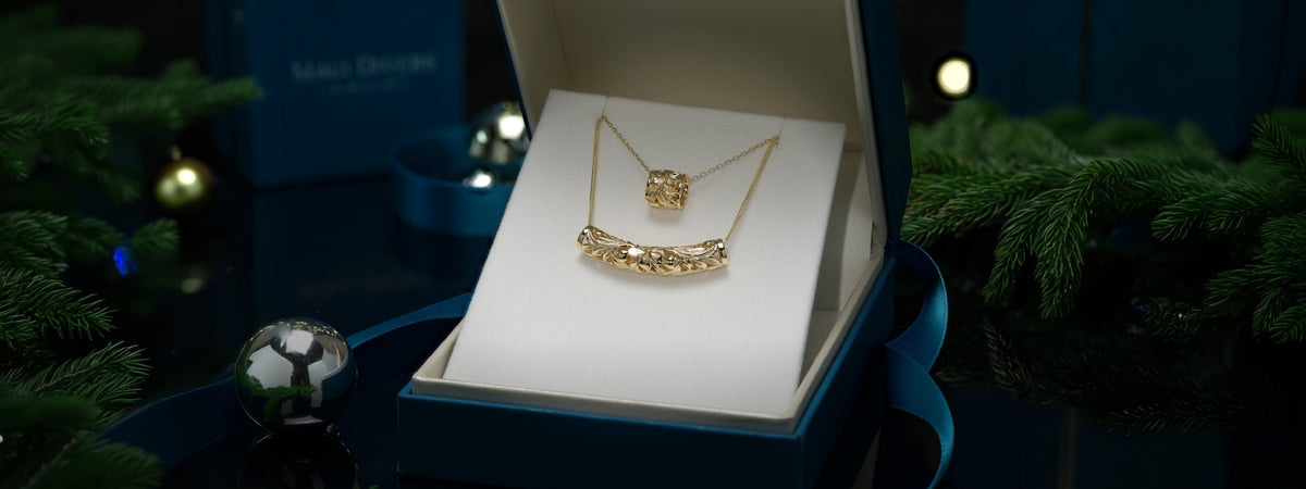 Two gold Hawaiian Heirloom necklaces displayed inside an open blue jewelry box on a white insert, surrounded by dark holiday greenery and ornaments.