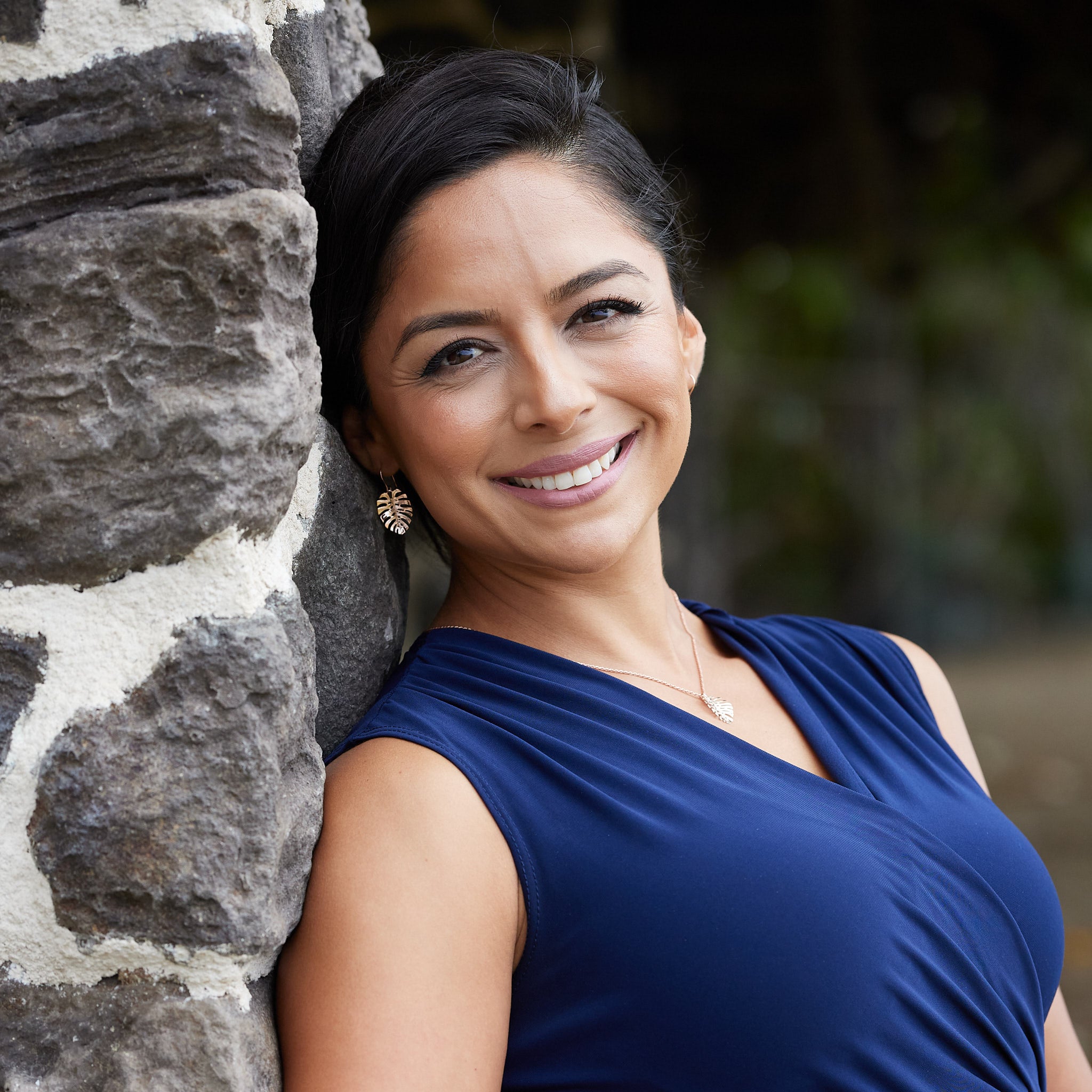 A woman leaning against a rock wall wearing a blue top and 23mm Monstera Earrings and Pendant in Rose Gold