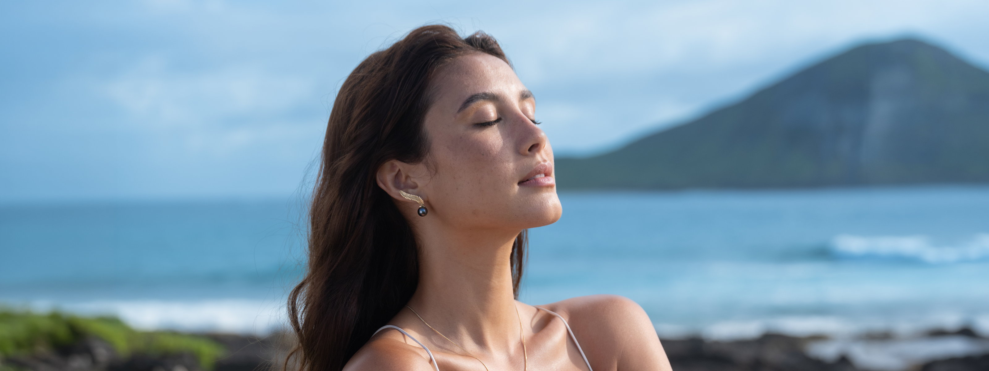 Woman with eyes closed standing and wearing a kupukupu fern Tahitian black pearl earring on a beach with mountains in the background