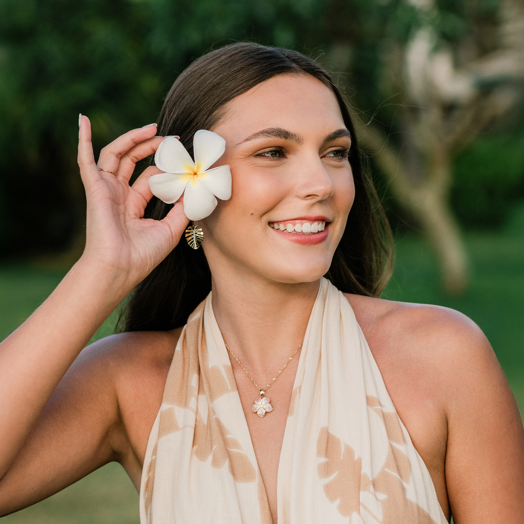 Woman placing a plumeria in her ear wearing a Plumeria Sapphire Pendant and Monstera Earrings in Gold with Diamonds