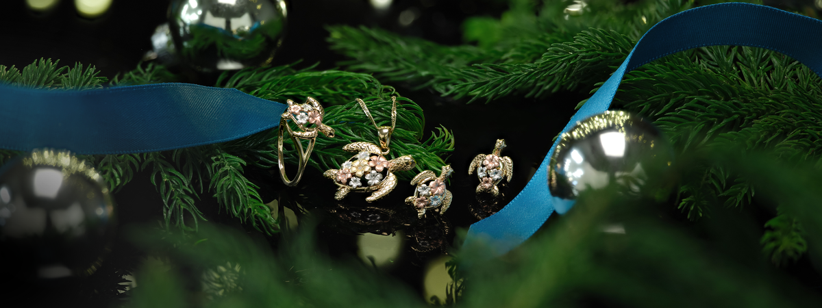 A set of 14k gold sea turtle pendant, ring, and earrings with plumerias on its back, set on a dark reflective surface with blue ribbons, silver ornaments, and pine leaves