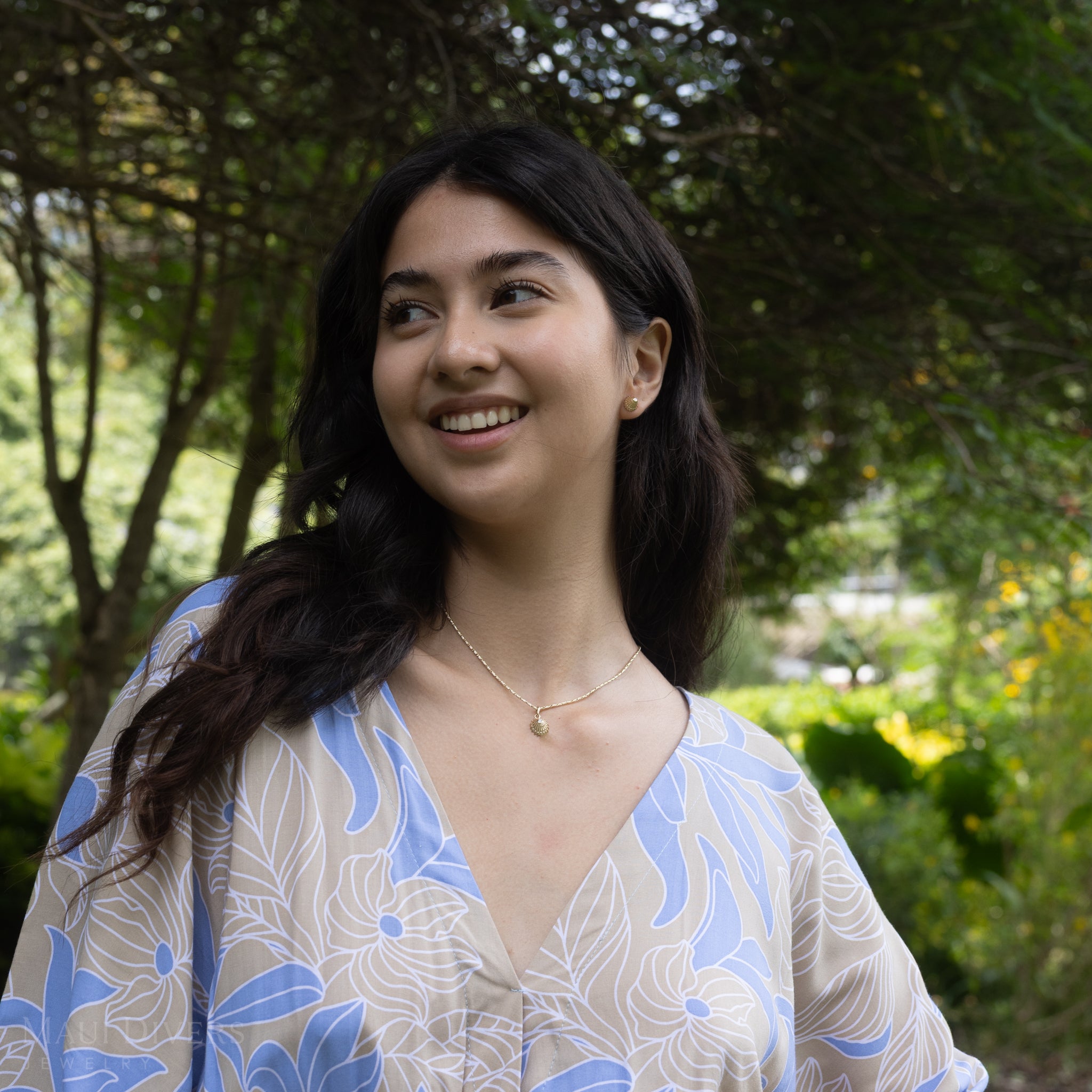 Woman wearing a gold necklace with an ʻOpihi diamond pendant against a green garden