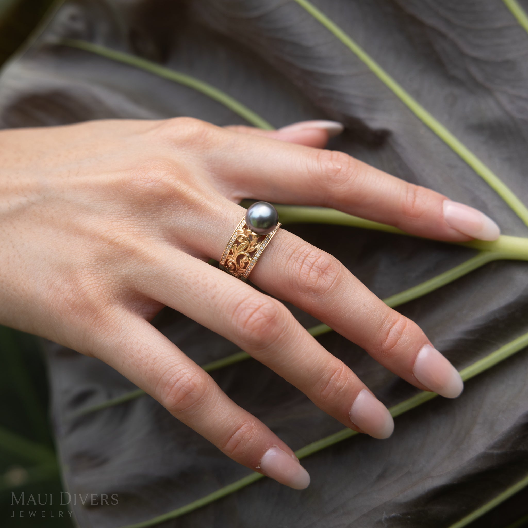 Hand wearing a Living Heirloom Tahitian Black Pearl Ring in Gold with Diamonds on a large green leaf