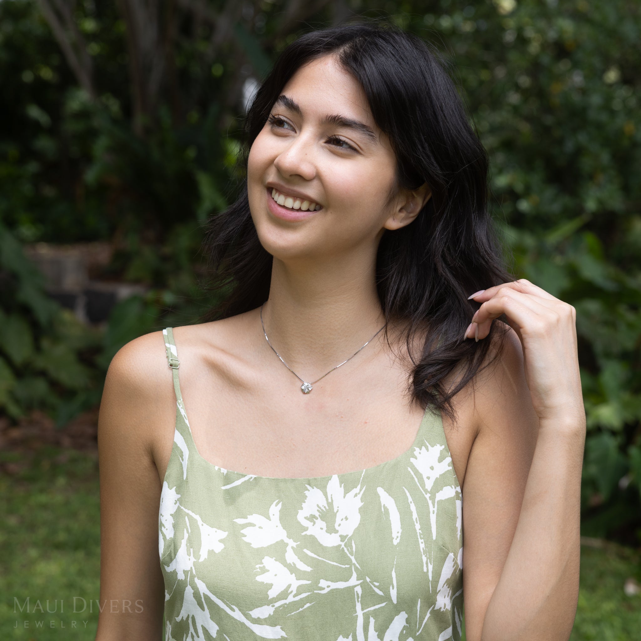 Woman in a green floral dress wearing a white gold hibiscus pendant with a blurry green background
